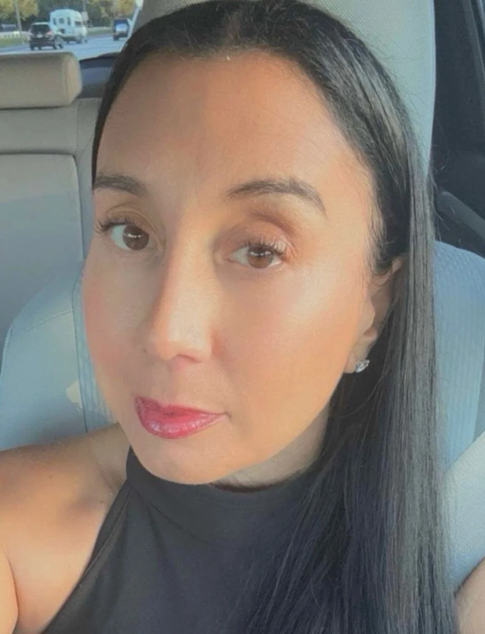 A woman with straight black hair, wearing makeup with red lipstick, and a sleeveless black top, sitting in a car with a highway visible through the window behind her.