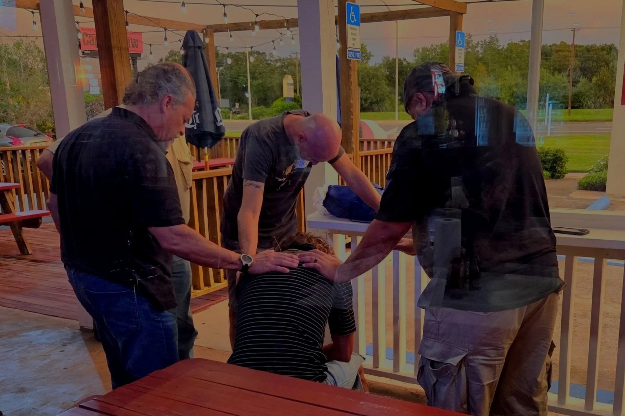 A group of people are gathered around a person who is kneeling and appears to be receiving medical attention at an outdoor venue with a wooden deck and string lights.