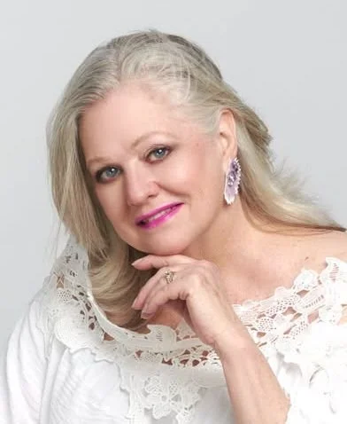 A mature woman with long gray hair, wearing a white lace top and large purple earrings, posing against a light background.