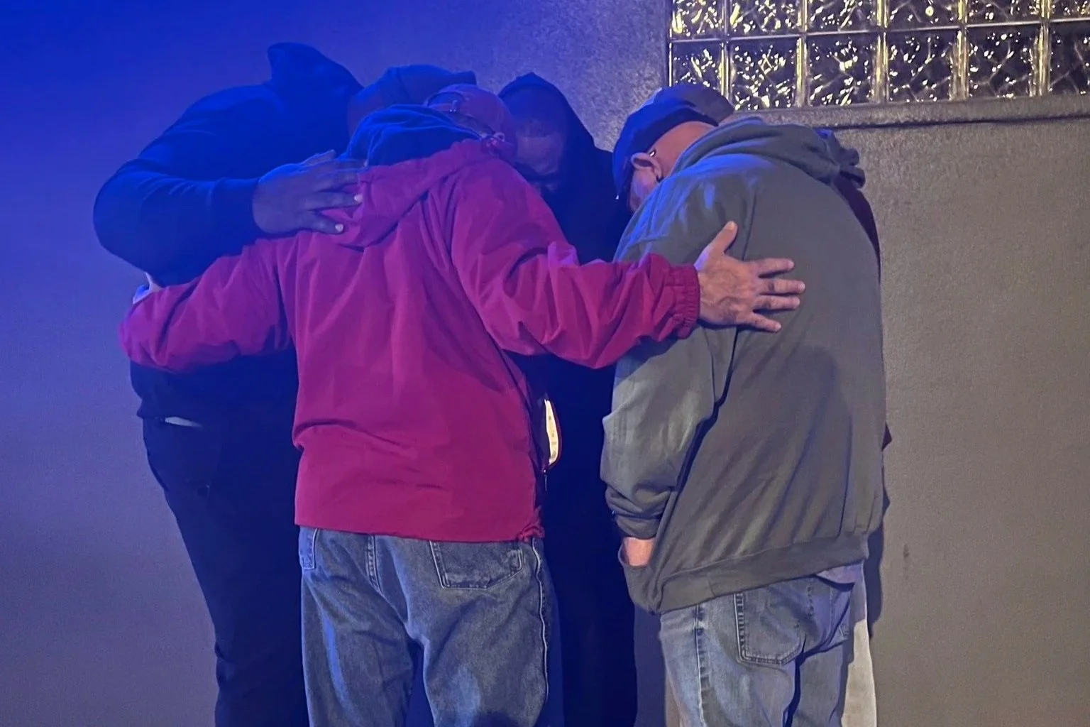 Three men are praying together, standing in a close circle with their heads bowed and hands on each other's shoulders, inside a building with a tiled wall and a light-colored wall.