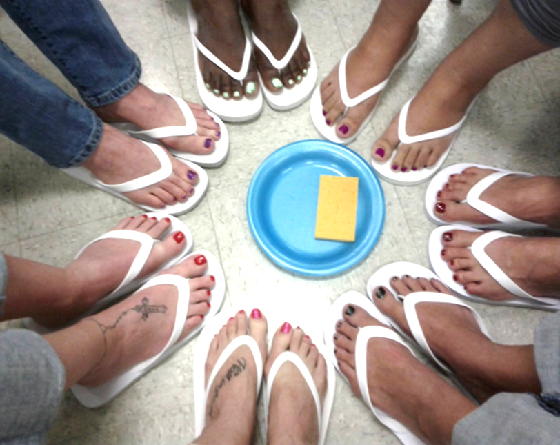 Group of people standing in a circle wearing white flip-flops, with a blue bowl and yellow sponge in the center, on a tile floor.