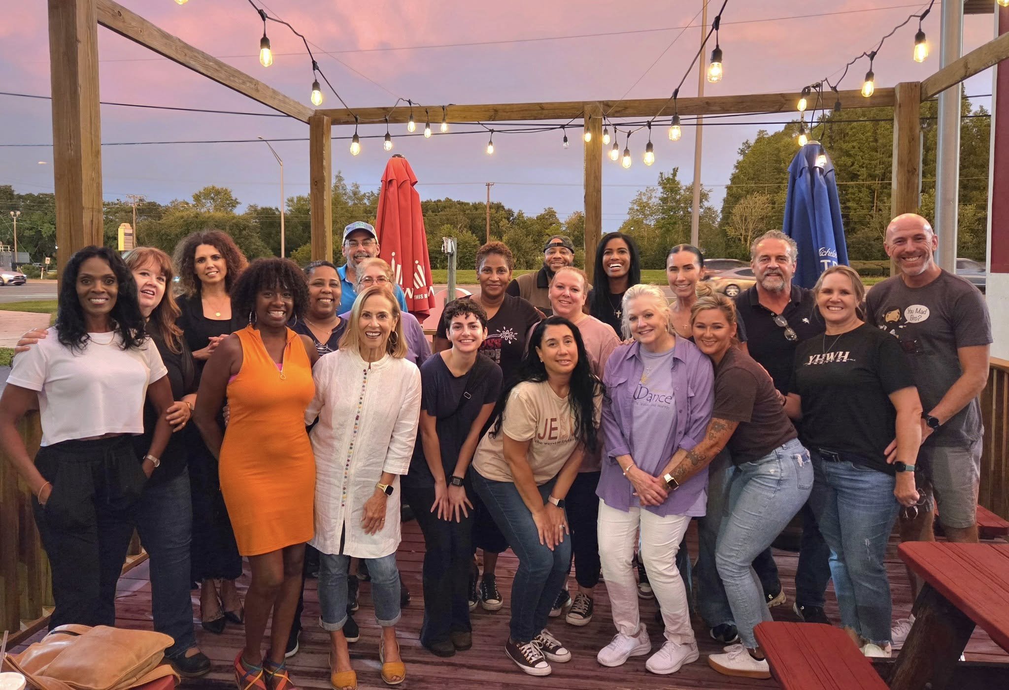 A group of people gathered together smiling on an outdoor deck with string lights overhead during sunset.