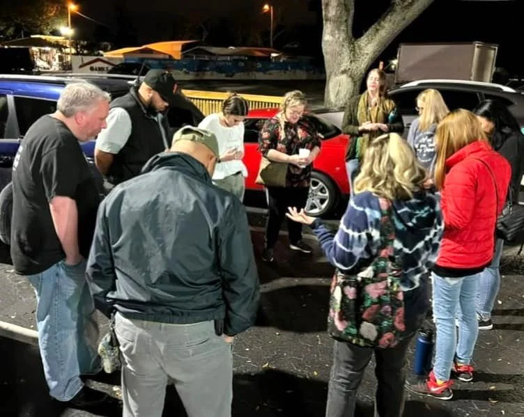 Group of people gathered in a parking lot at night with a red vehicle and a tree in the background, some holding papers and engaged in conversation.