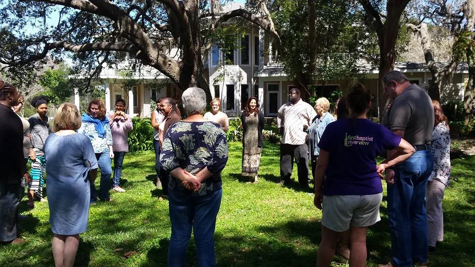 A group of people standing outdoors in a circle on a grassy yard, under large trees, with a house in the background, participating in a gathering or event.