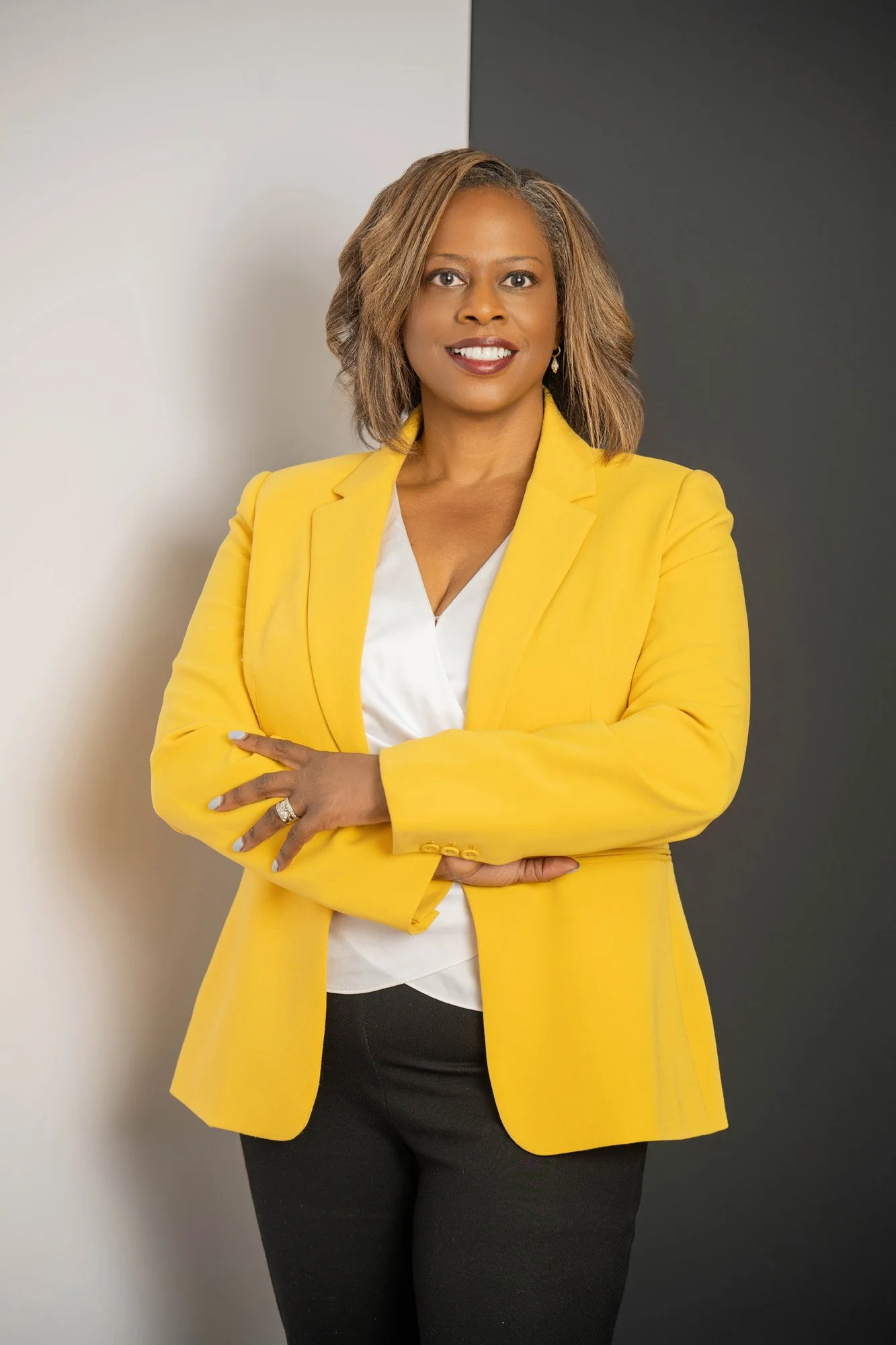 A woman with shoulder-length hair wearing a yellow blazer over a white top, standing against a two-tone gray and white background, smiling with her arms crossed.