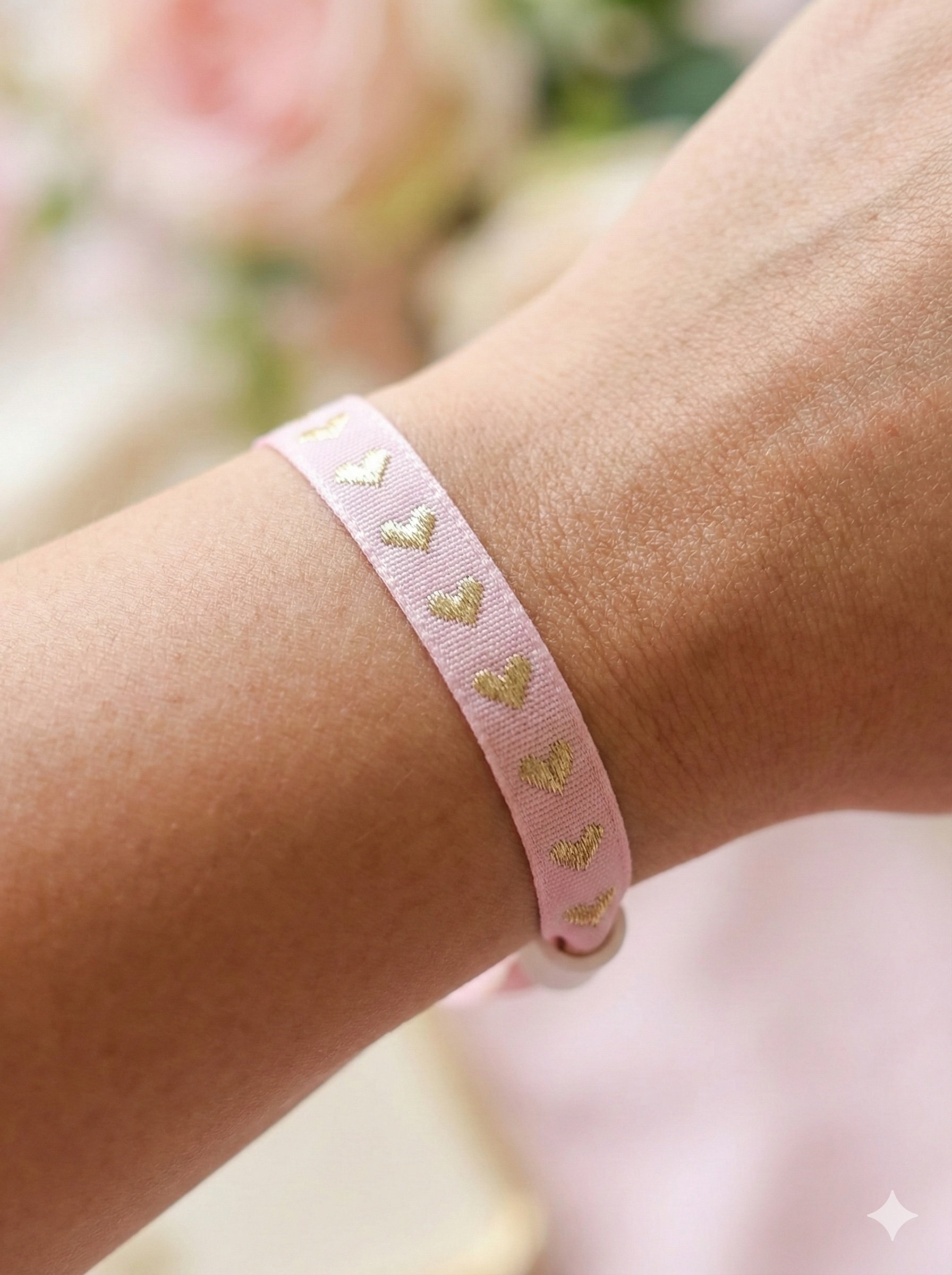 pastel pink satin ribbon bracelet on a wrist featuring embroidered gold hearts and a toggle clasp.