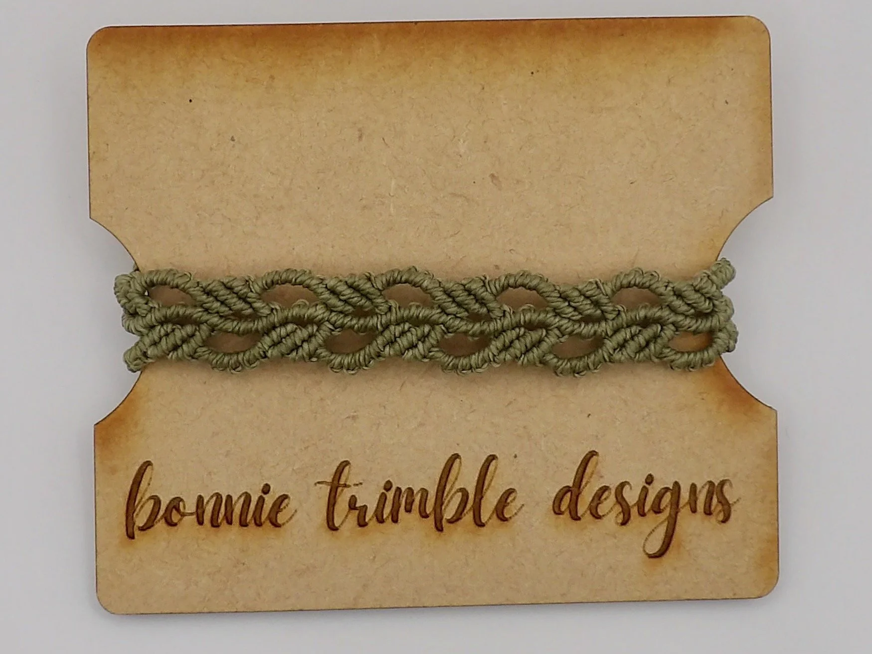 A person wearing a delicate green micromacrame ivy leaf bracelet, showcasing how it wraps around the wrist against a soft, out-of-focus natural background.