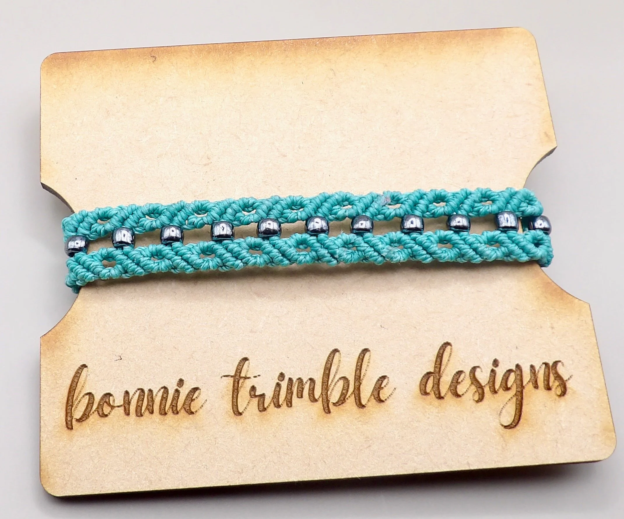 Handmade teal micromacrame bracelet with dark gray seed beads on a white background.