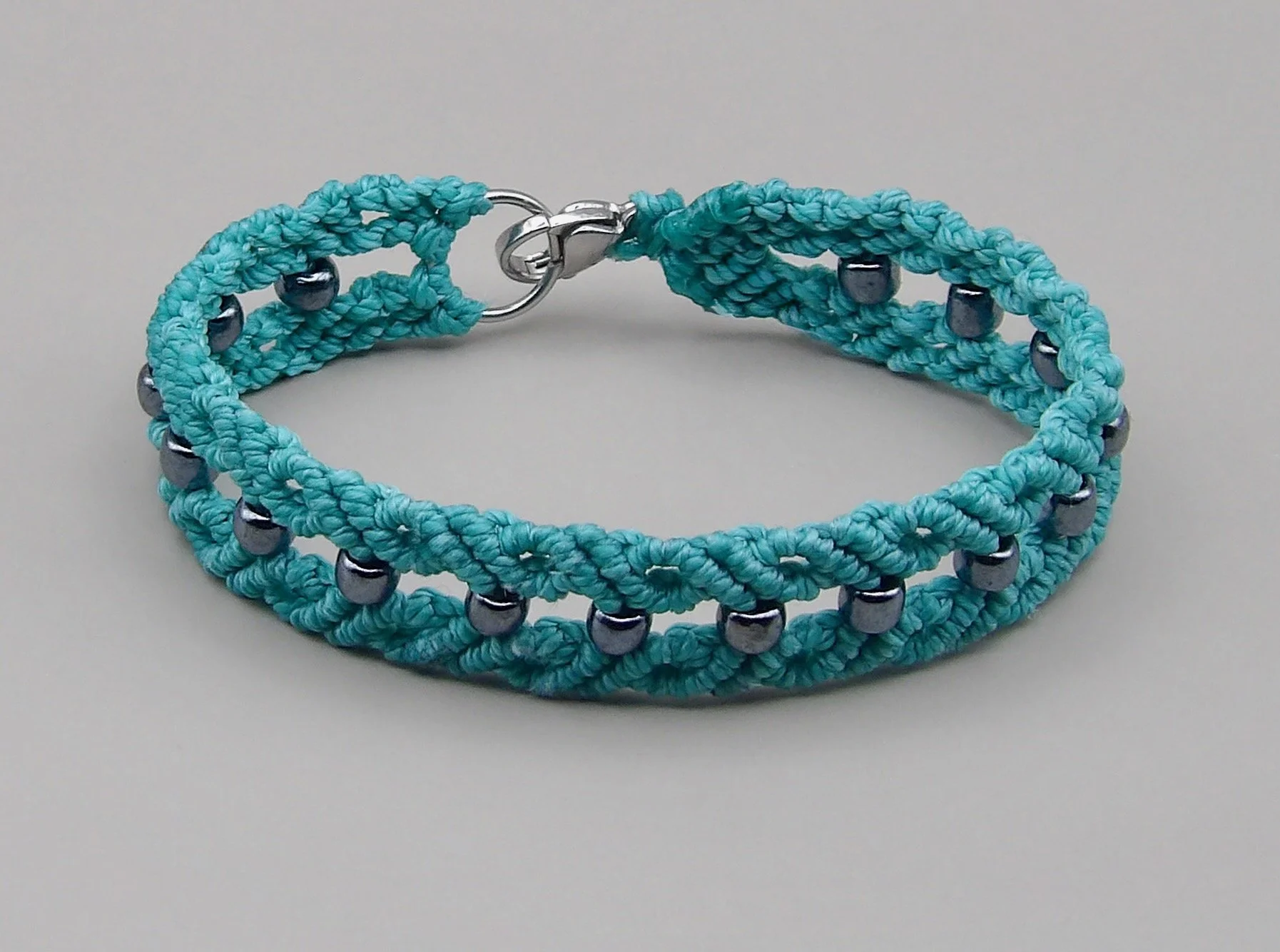 A close-up of a hand-woven teal macrame bracelet, featuring a pattern of small, silver-lined dark gray glass beads, and a stainless steel lobster clasp.