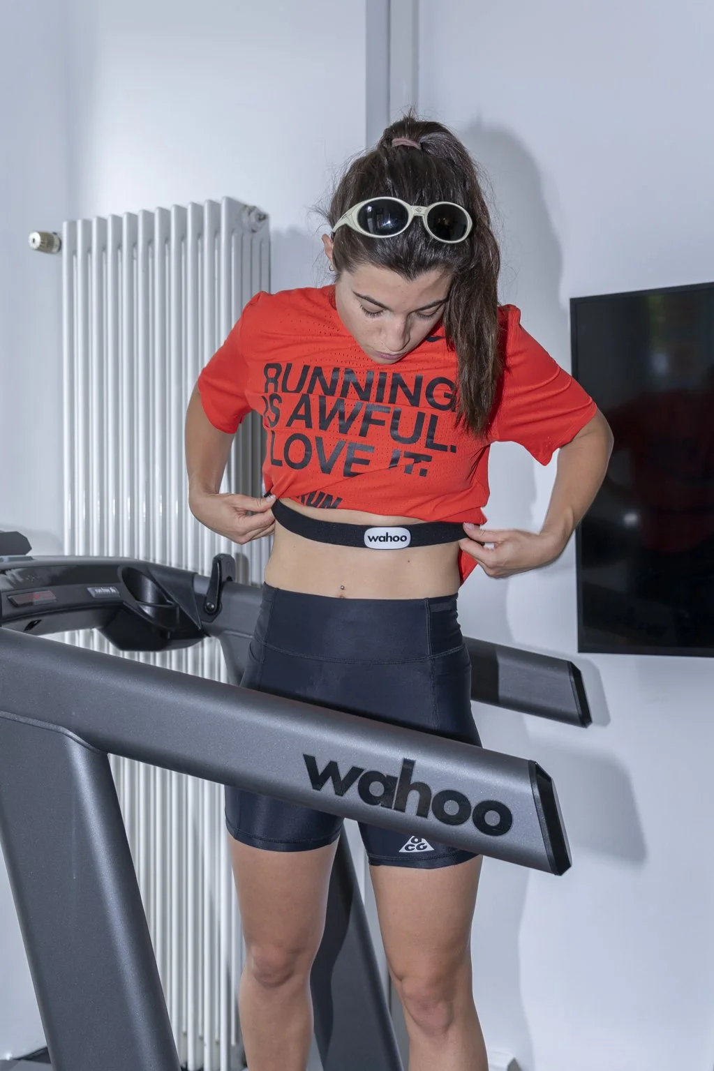 Junge Frau beim Kleidung wechseln auf einem Laufband, trägt eine rote T-Shirt mit schwarzen Schriftzug und schwarze Sporthose, Sonnenbrille auf dem Kopf, Fitnessstudio-Umgebung.