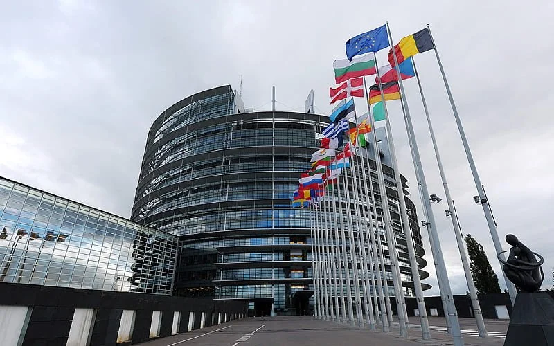 Modern glass building with multiple floors and rounded architecture, displayed with numerous flags from different countries on tall poles in front of it, on a cloudy day.