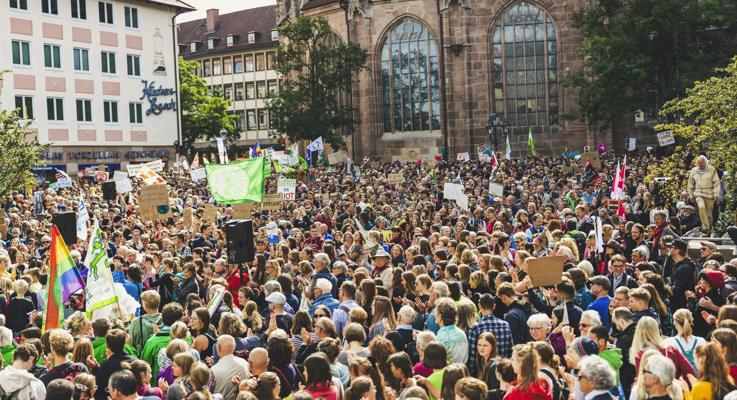 Crowd of people protesting with signs and flags near a church with large stained glass windows.