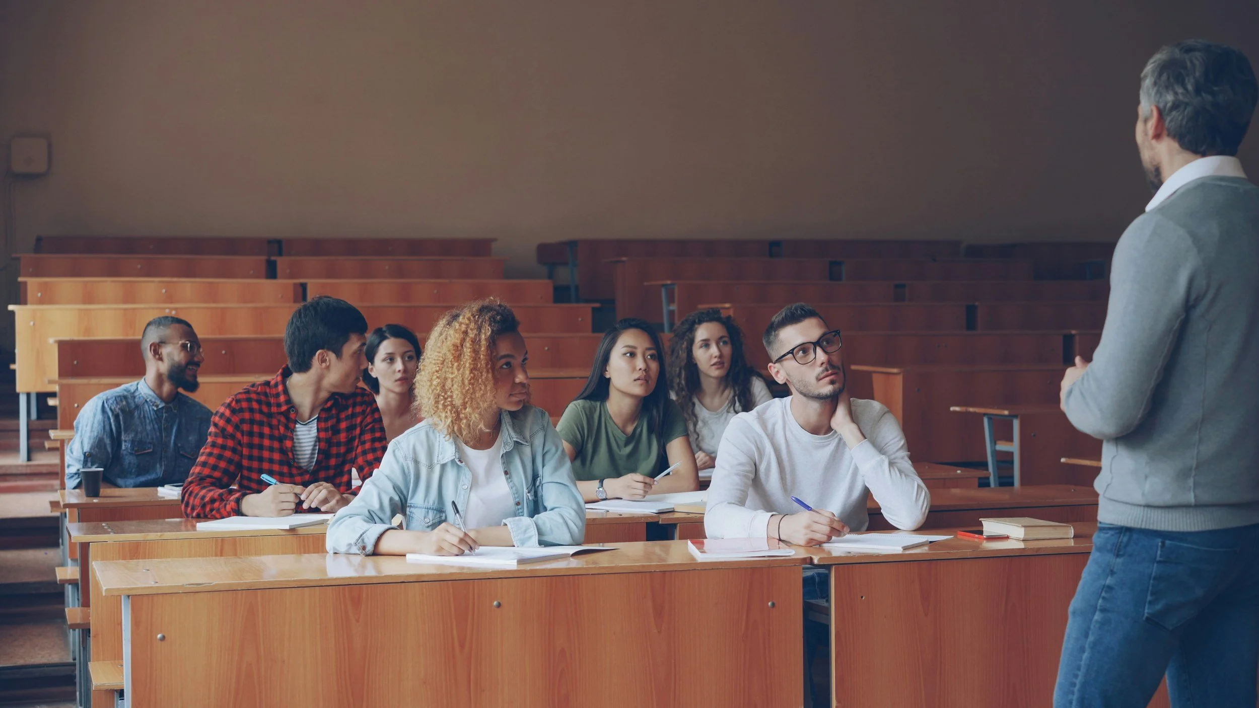 A university classroom with students sitting at desks while a male professor stands at the front of the room.