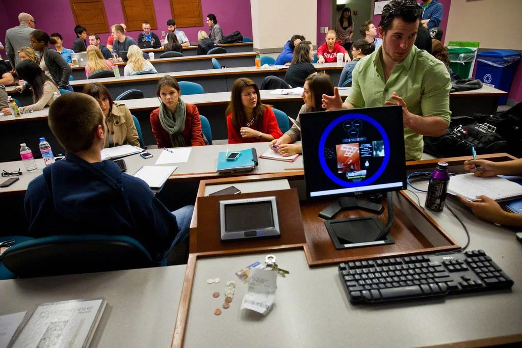 A group of students sitting around a classroom desk engaging in a discussion with a man standing and speaking to them. The classroom has purple walls, desks with computers, notebooks, and bottles of water.