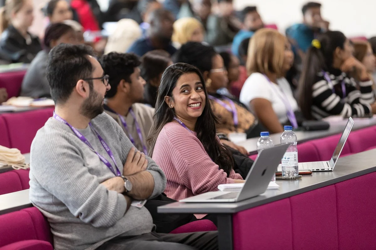 A diverse group of students attending a lecture or conference, sitting in rows with laptops and water bottles, paying attention and engaging.