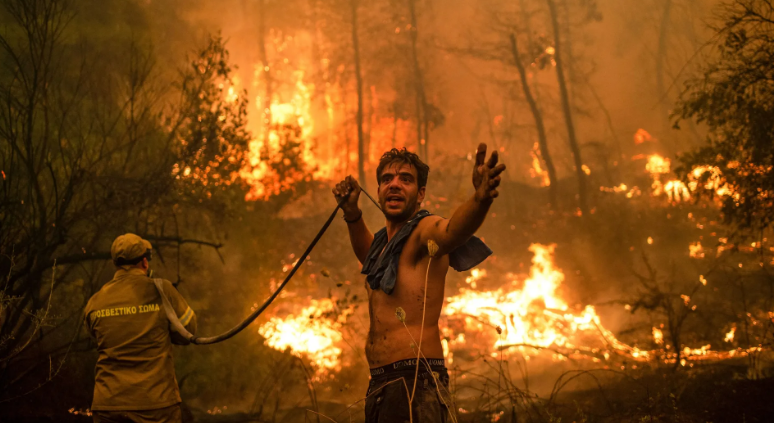 A shirtless man with a beard and dark hair gestures with his arms while a firefighter in a yellow uniform faces away, holding a hose, in a forest fire scene.
