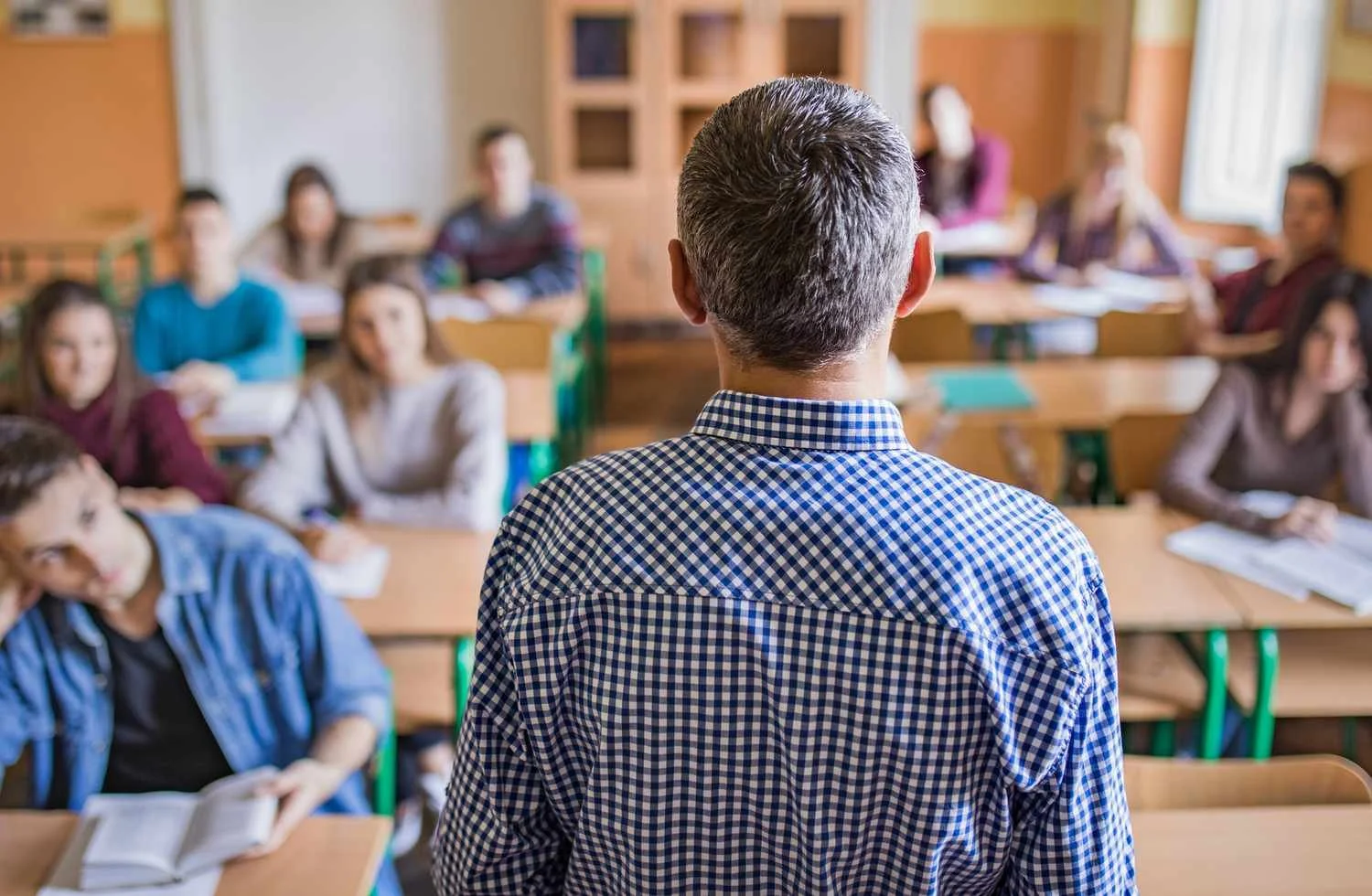 A teacher standing in front of a classroom of students, viewed from behind, with students at desks looking at the teacher.