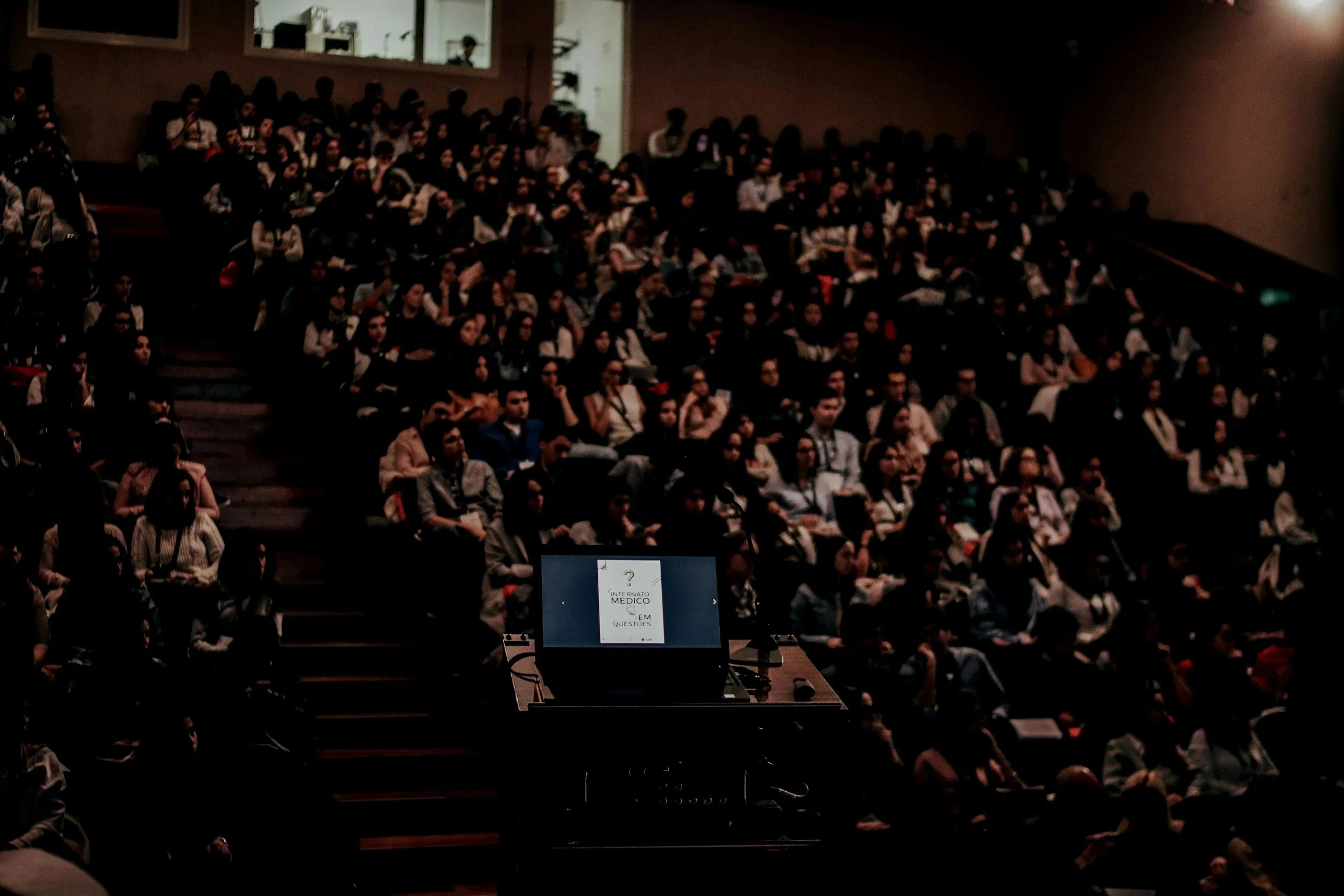 A large audience seated in an auditorium, watching a presentation at a conference or seminar, with a projector screen displaying some text.