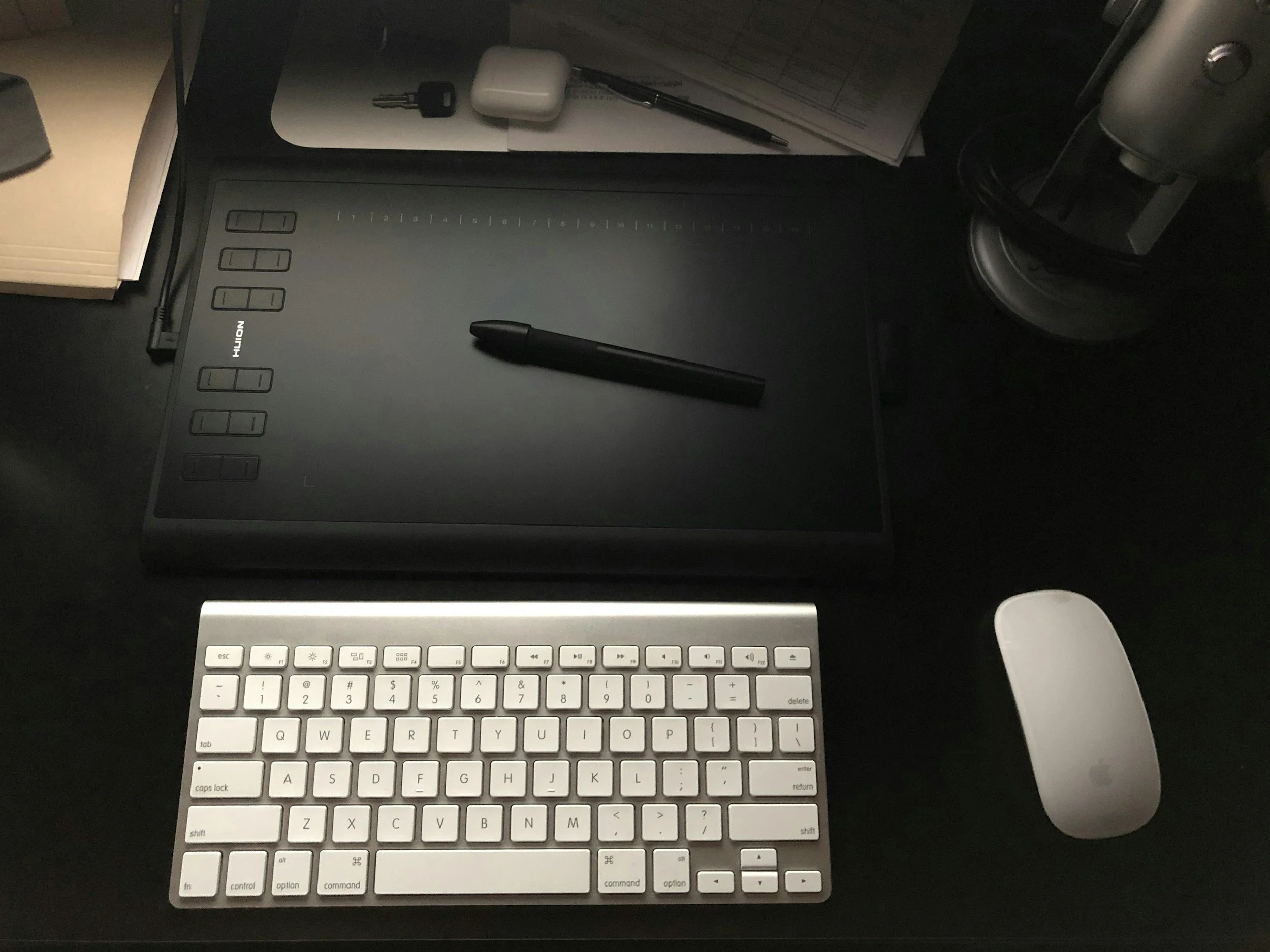 A workspace featuring a graphics tablet, a wireless keyboard, a white mouse, a pen, a notebook, a key, and earbud case on a dark desk.