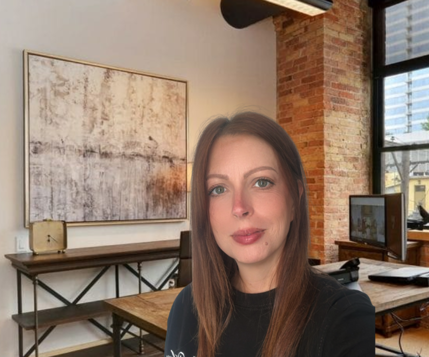 A woman with long brown hair and green eyes taking a selfie in an industrial-style office with exposed brick walls and large windows.