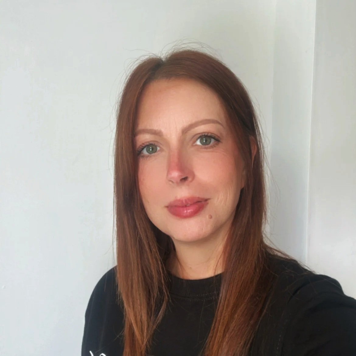 Close-up of a woman with long reddish-brown hair, light skin, and green eyes, wearing a black shirt, standing against a plain light-colored background.