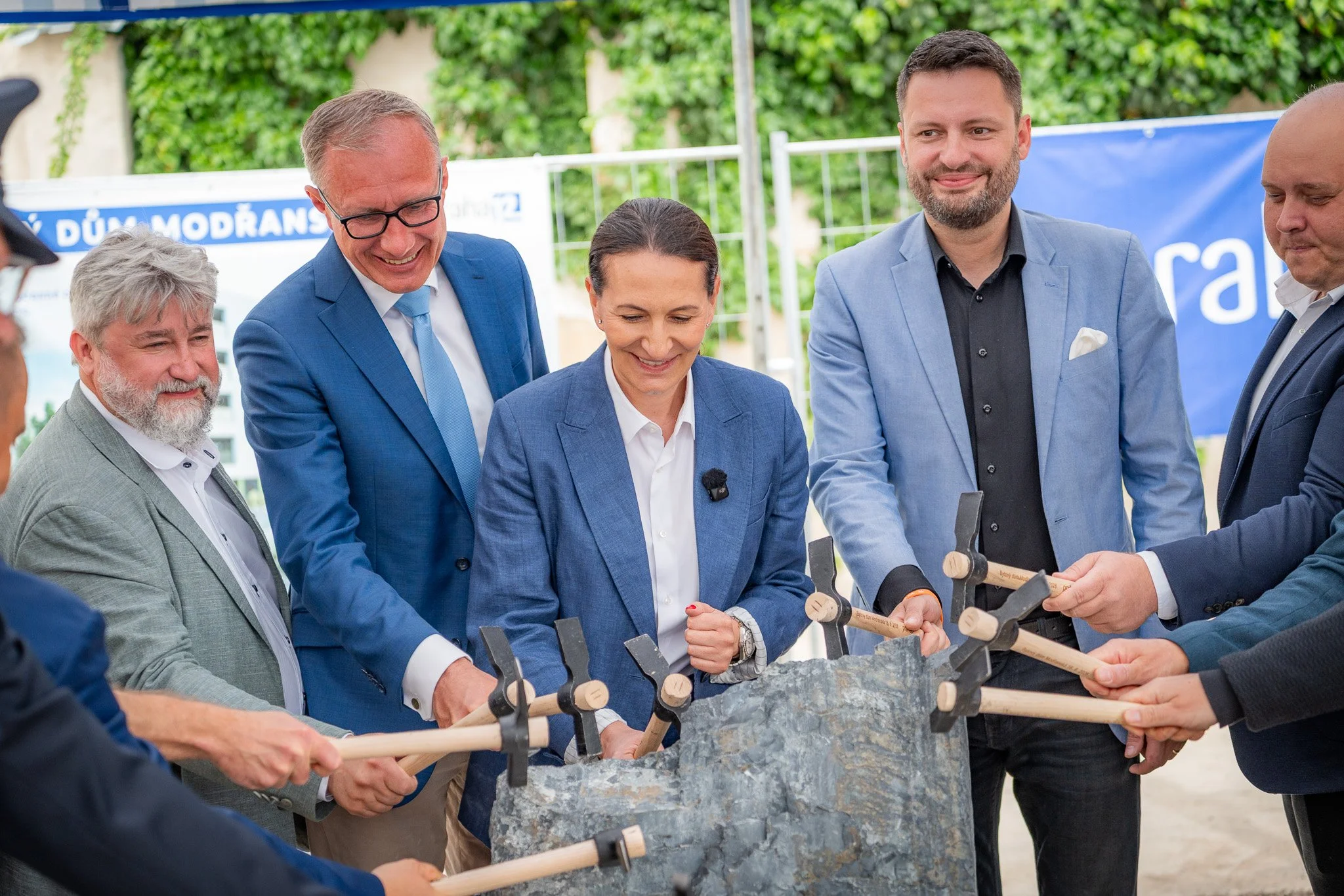 Group of professionally dressed people participating in a groundbreaking ceremony, holding shovels with wooden handles and shoveling dirt.