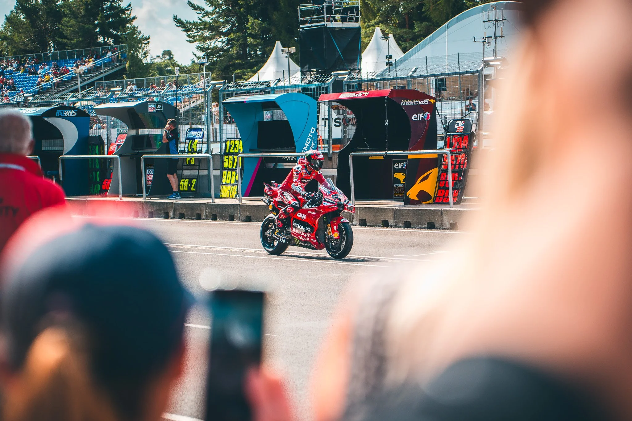 Race car driver on a red motorcycle, preparing for a race at a racing circuit, with pit stop and grandstands in the background, seen through blurred foreground objects.