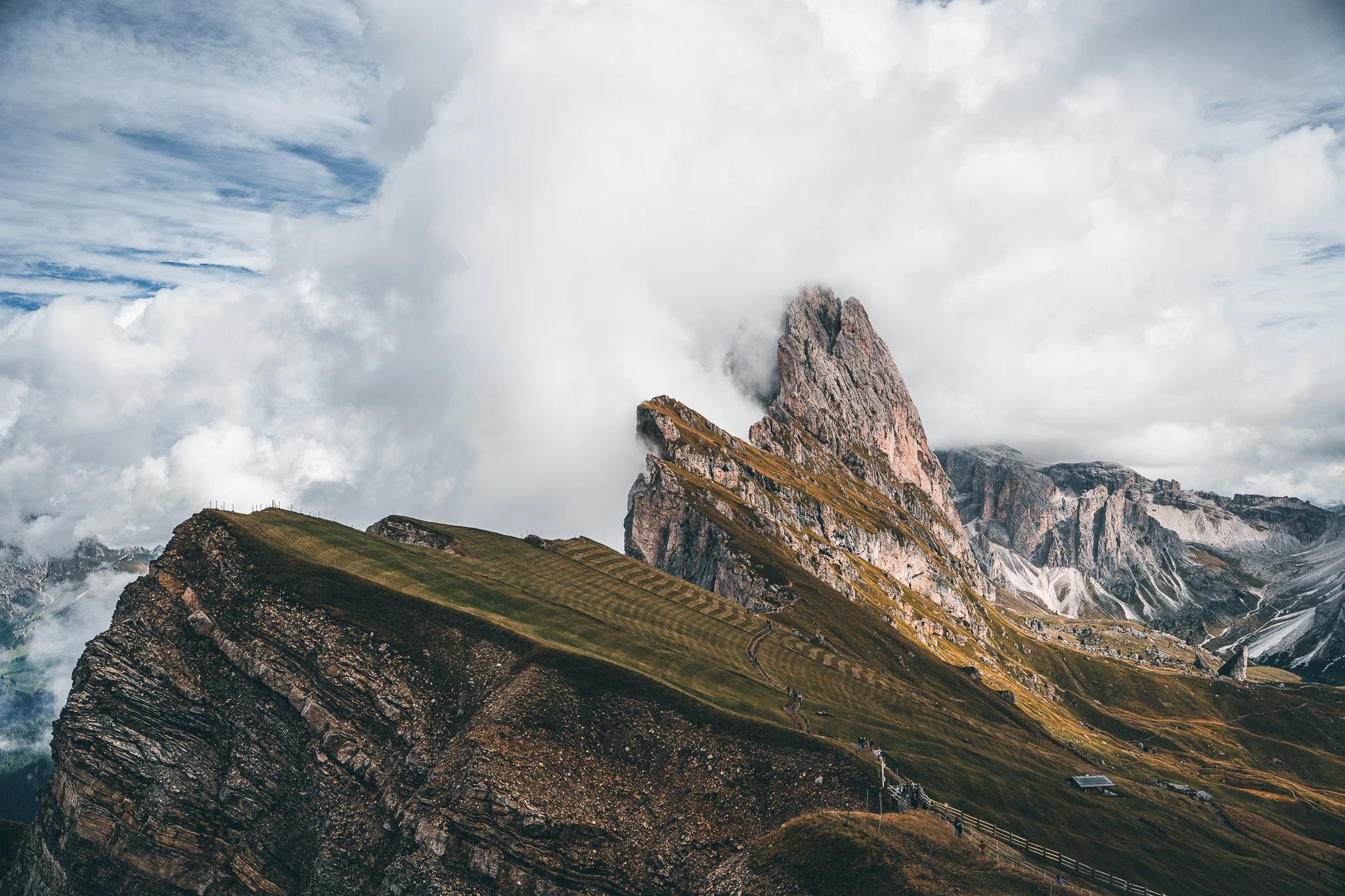 Mountain landscape with rocky peaks, green terraced slopes, and a cloudy sky.