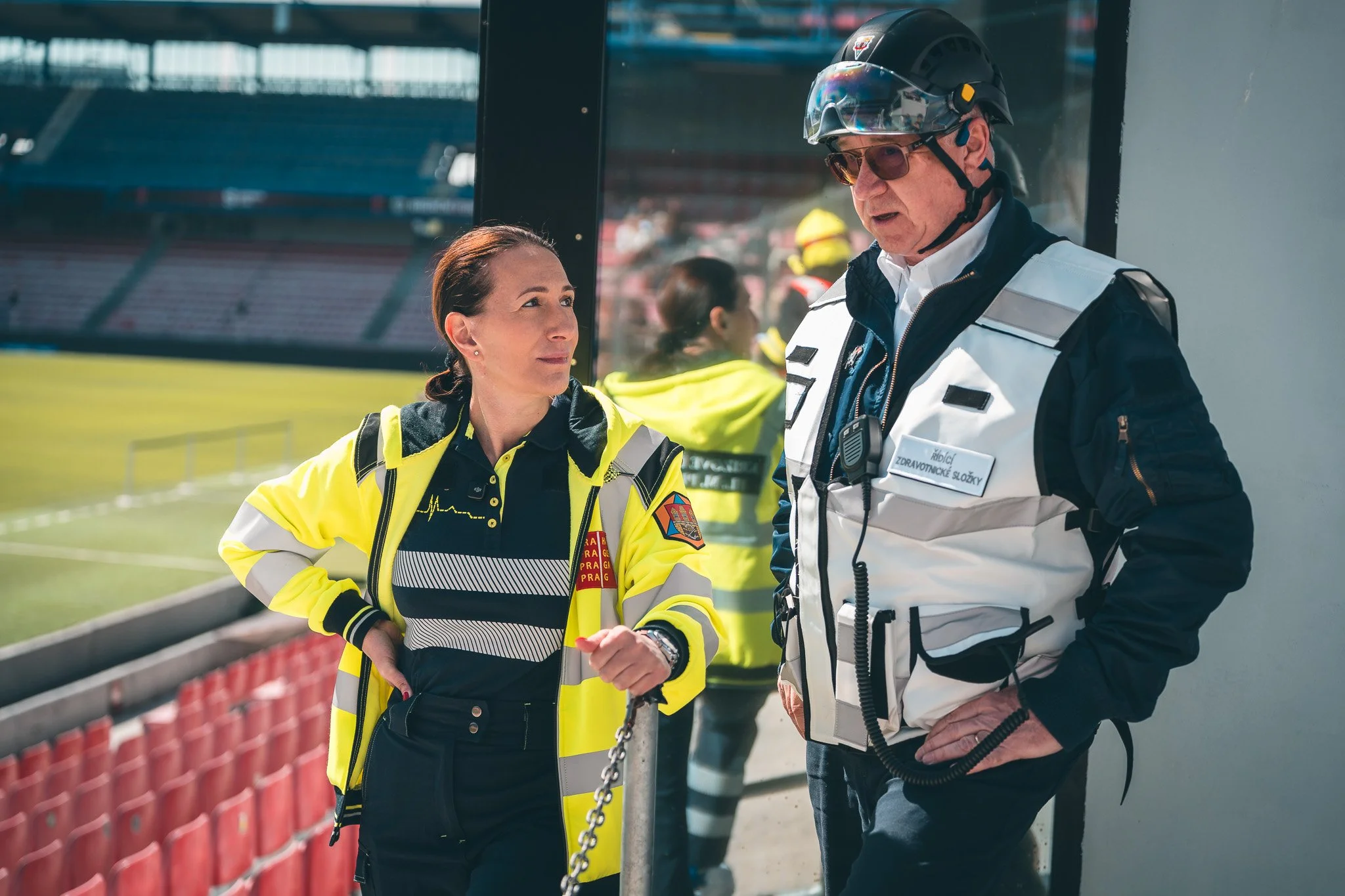 A woman and a man are having a conversation at a sports stadium. The woman is wearing a yellow safety jacket with emergency service insignia, and the man is wearing a white emergency service vest and a helmet with a visor. They are standing near a ba