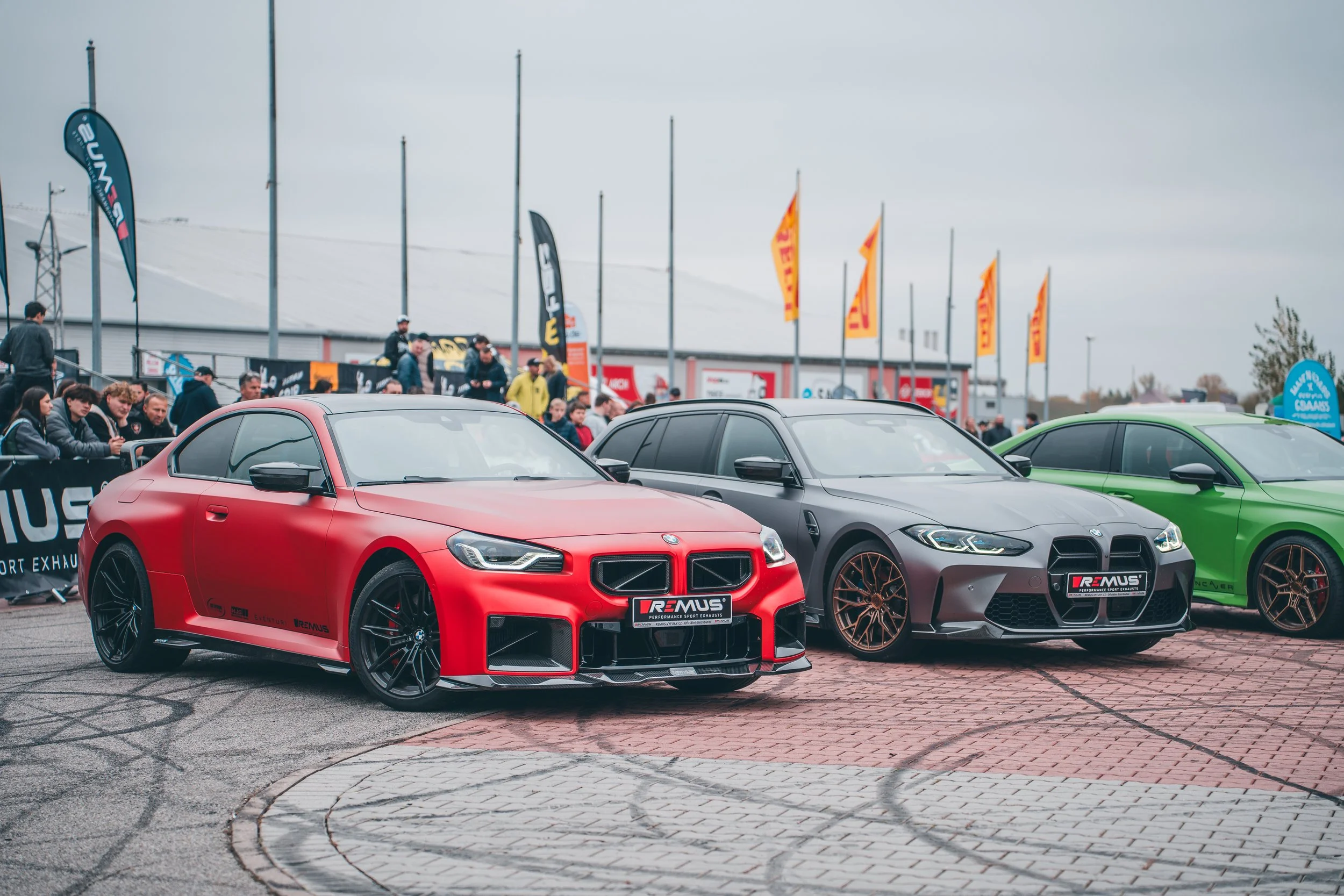 Several modified cars are parked at an outdoor car event, with a crowd of people in the background and flags fluttering in the wind.