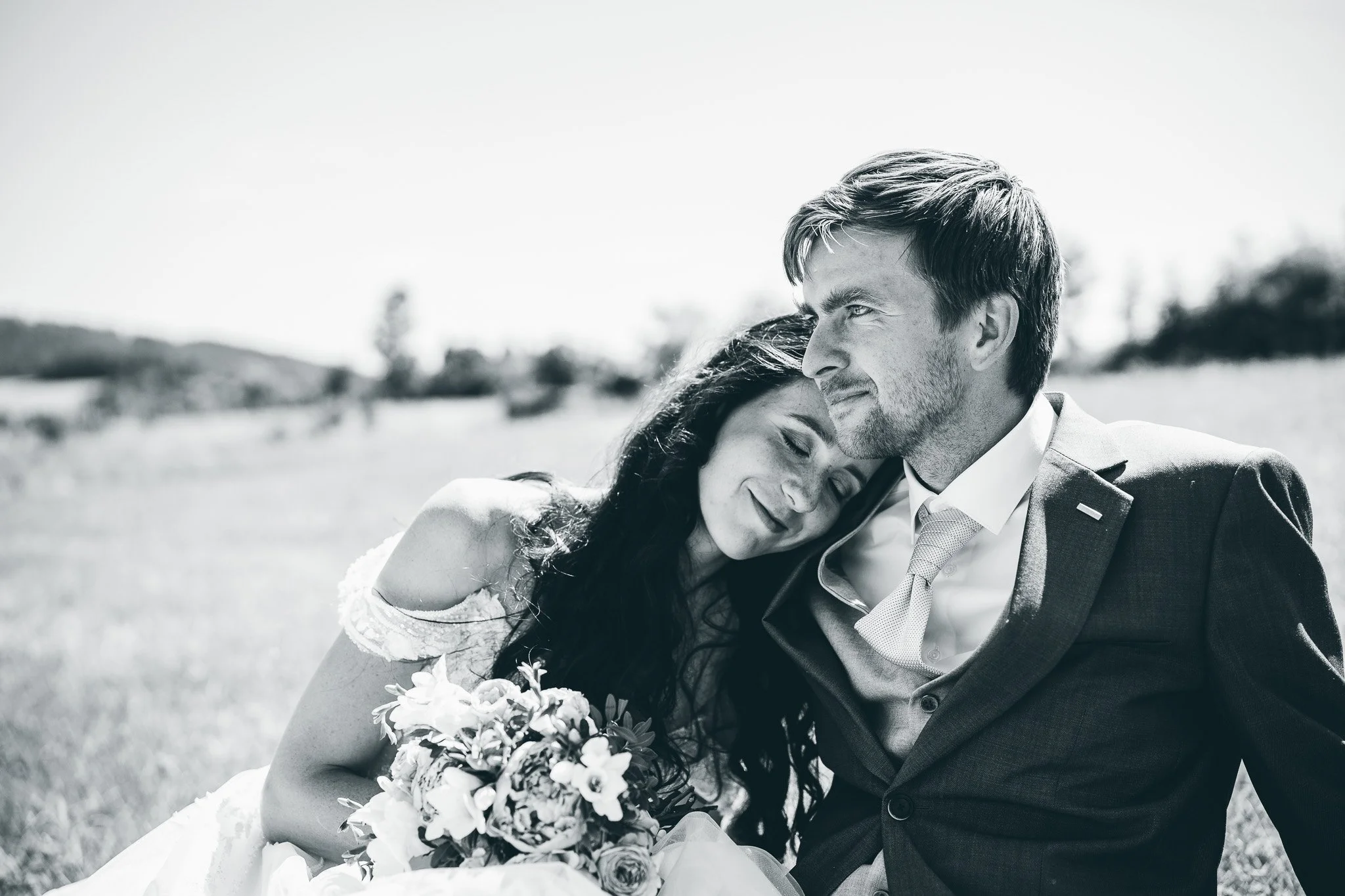 A black and white photograph of a couple with closed eyes, embracing outdoors. The woman is resting her head on the man's shoulder, holding a bouquet of flowers, with a blurred landscape in the background.