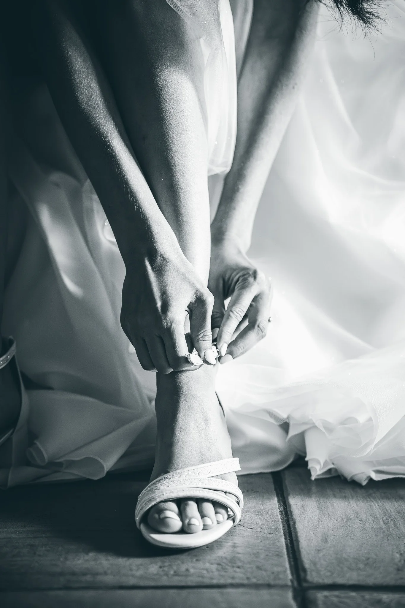 A close-up of a person tying the laces on their sandal while sitting on a tiled floor, with white fabric around them and sunlight creating shadows.