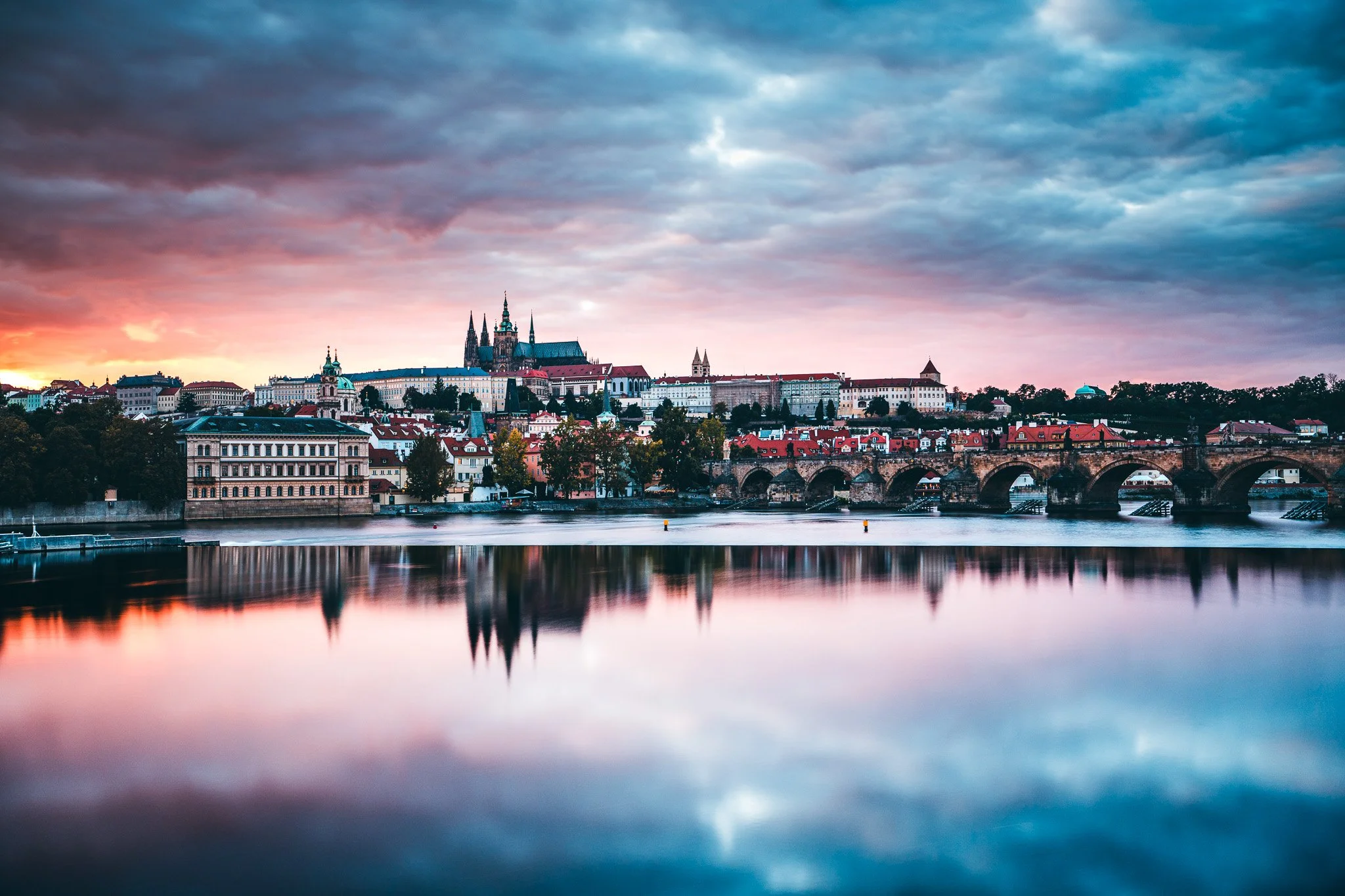 A cityscape view at sunset with historic buildings, a castle or cathedral, and a river reflecting the sky and city lights.