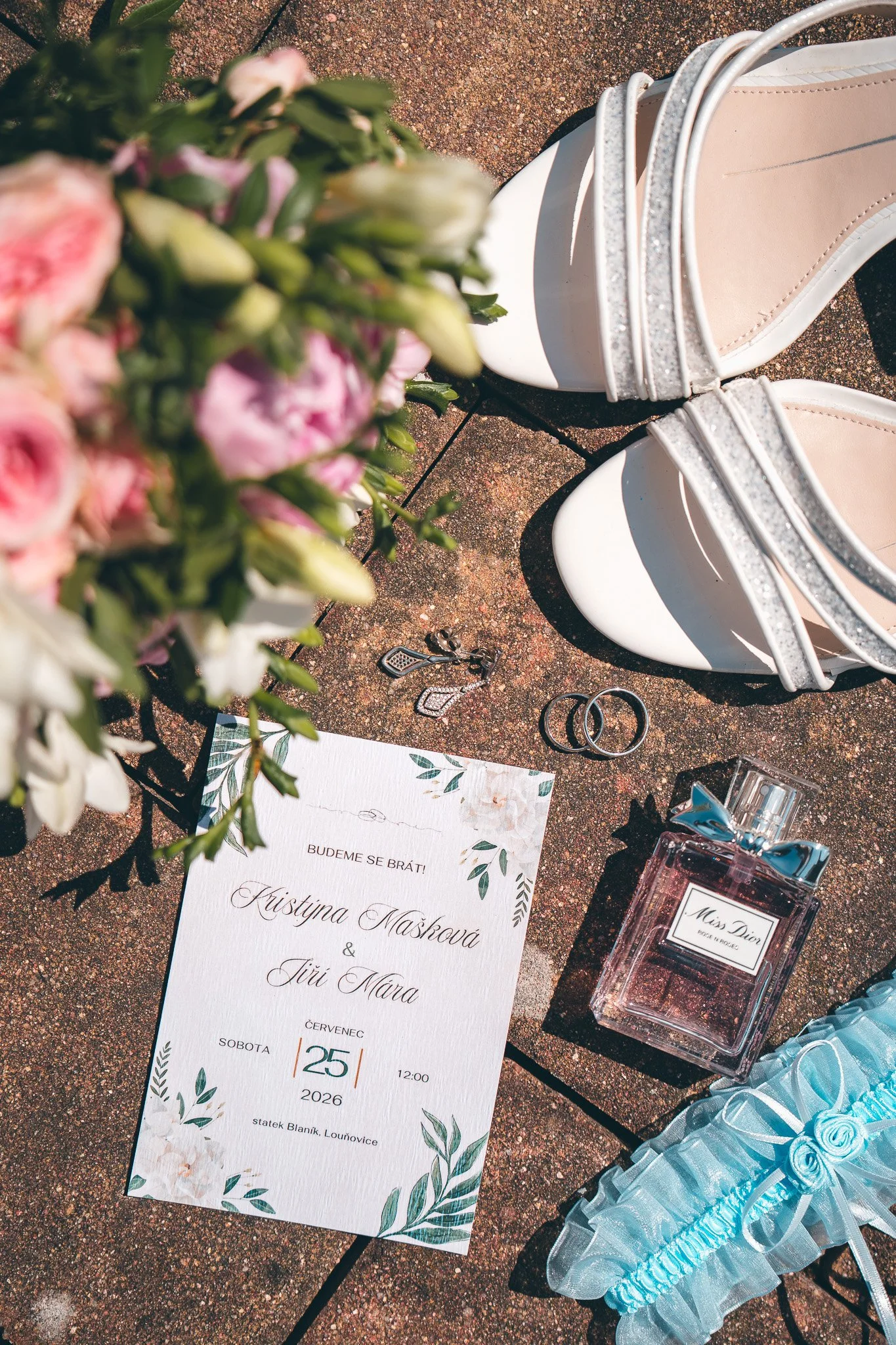 A wedding invitation card with floral design, lying on a textured surface, surrounded by flowers, rings, perfume, and blue ribbon decorations.