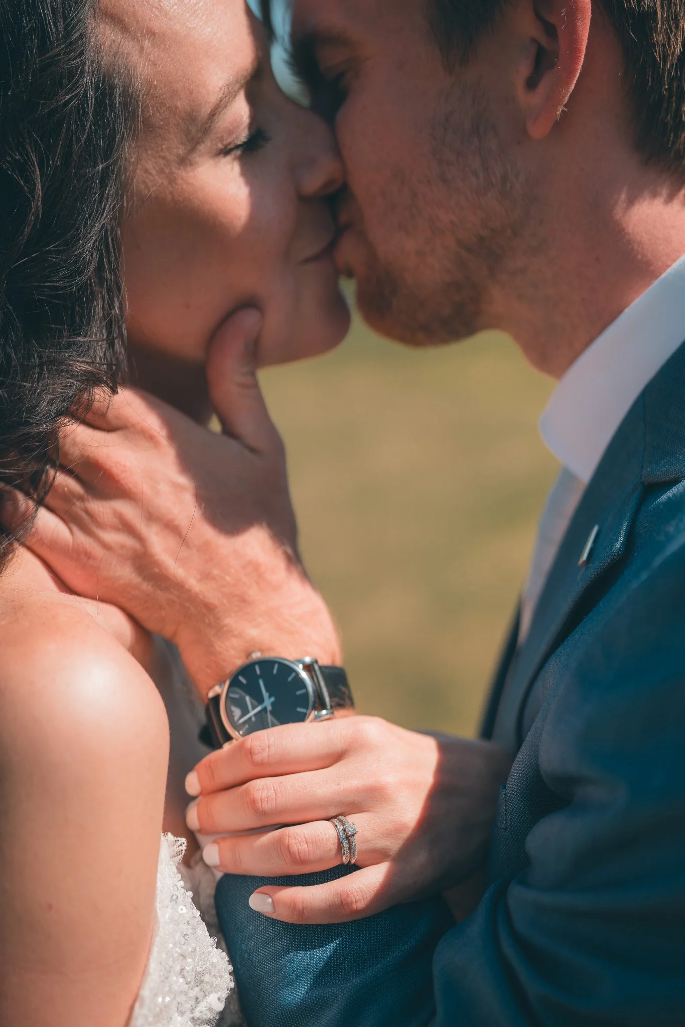 A close-up of a couple kissing outdoors. The woman has a lace dress and a watch on her wrist, and the man is wearing a suit. They are holding each other, and the woman's hand, showing rings, rests on the man's chest.