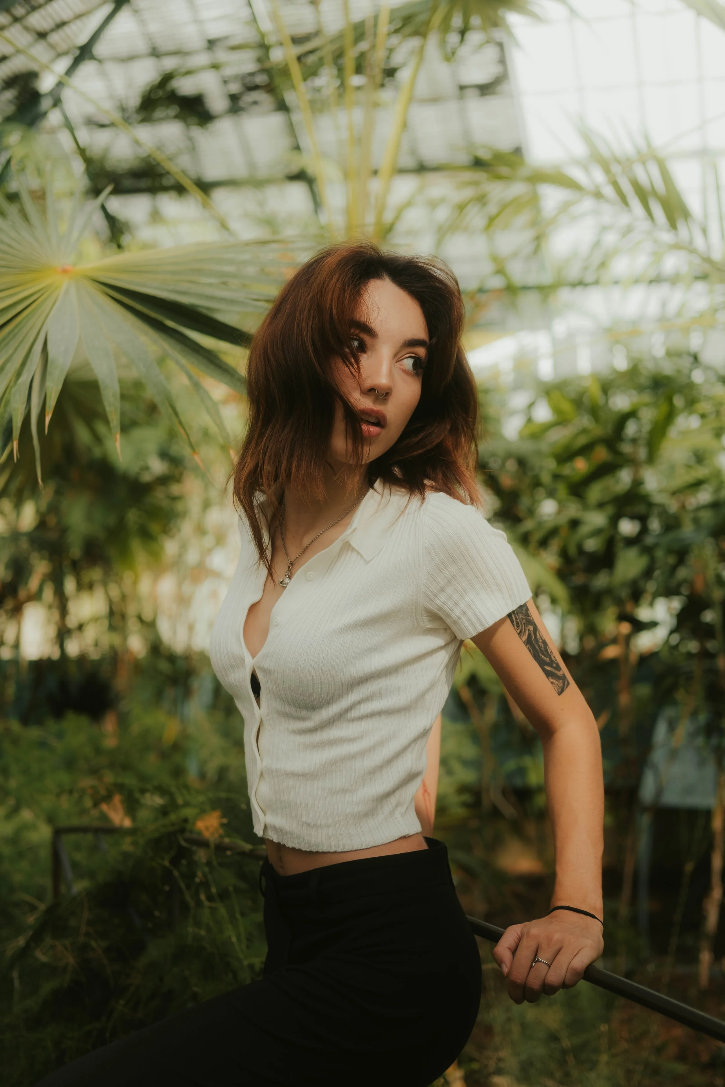 Jovem mulher com cabelos castanhos e tatuagens, usando blusa branca e calças pretas, posando em um ambiente de jardim com plantas tropicais.