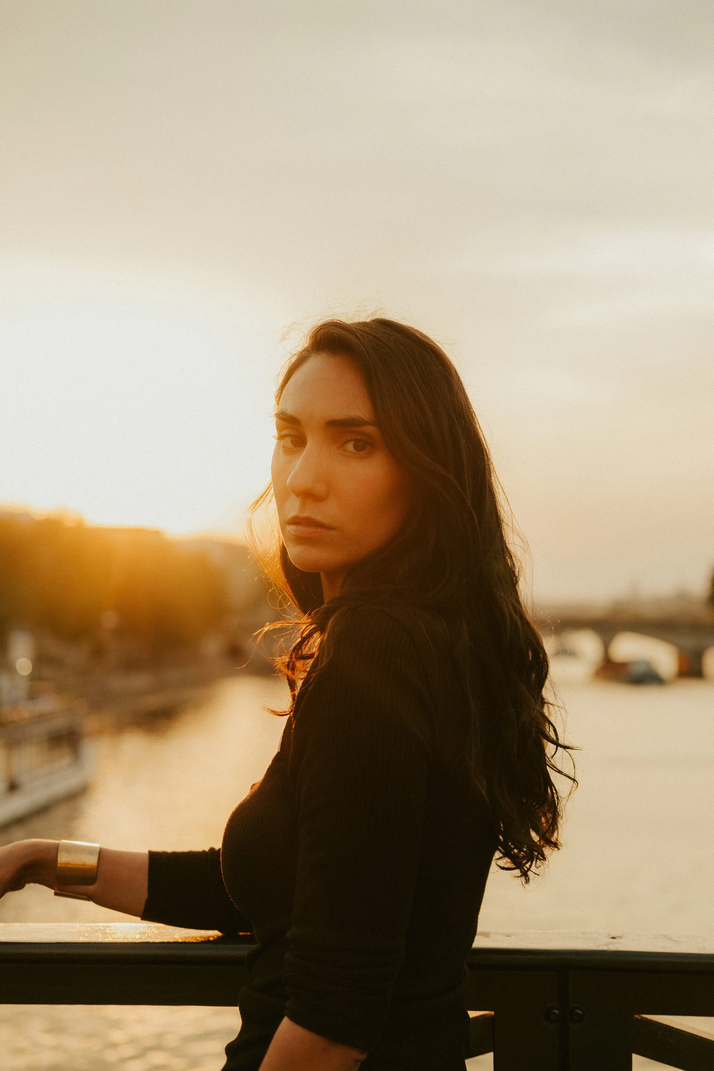 Mulher com cabelo castanho escuro e roupas pretas, de cabelo longo, posa ao atardecer perto de um rio.