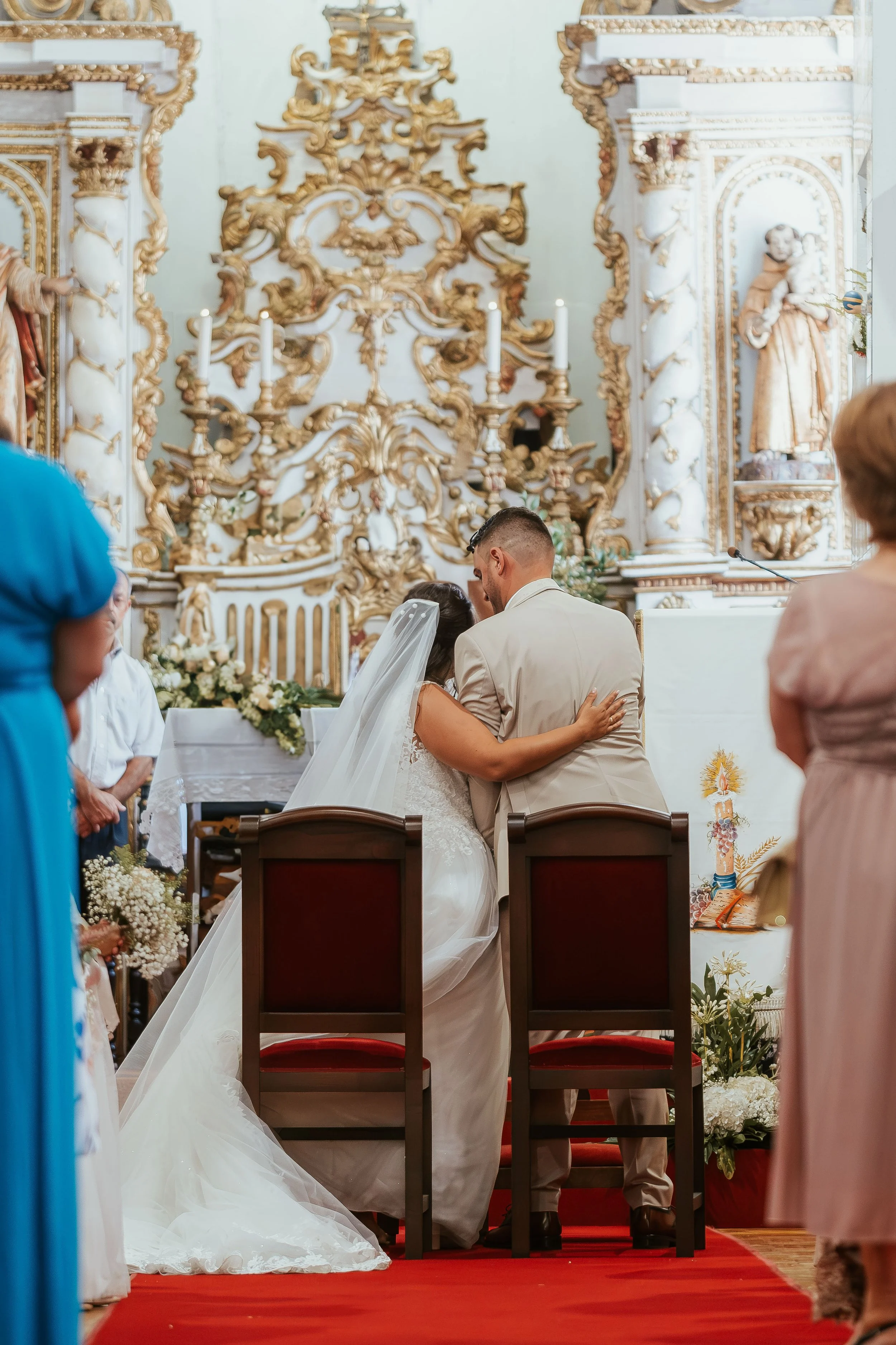 Casal de noivos se beijando no altar de uma igreja decorada com ornamentos dourados e brancos, durante a cerimônia de casamento.