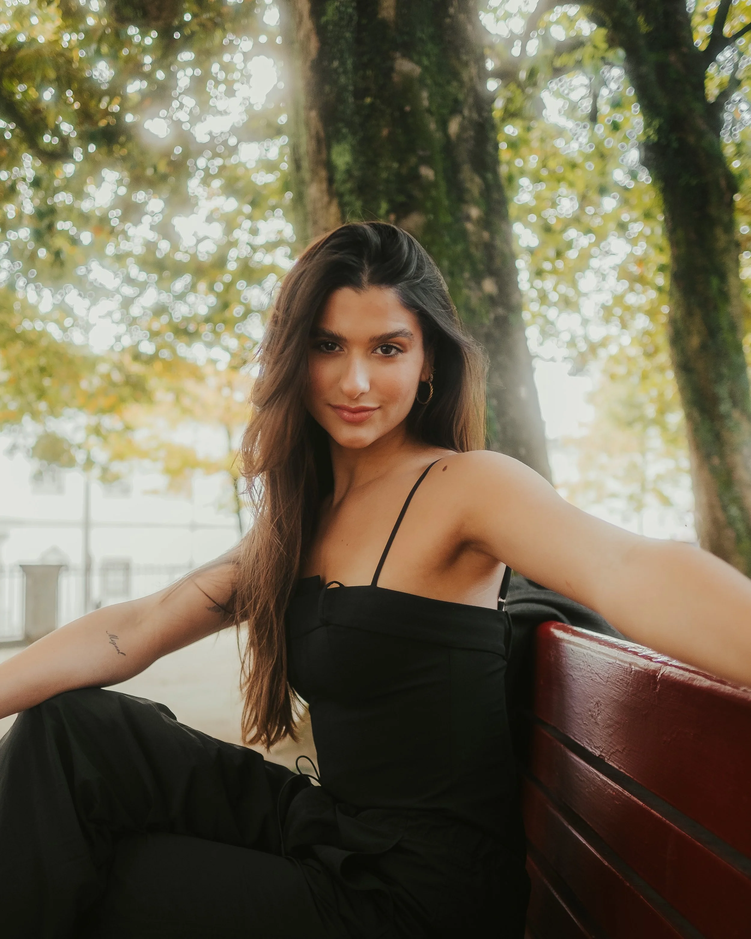 Jovem mulher com cabelo castanho, usando vestido preto de alças finas, sentado em um banco de parque sob árvores com folhas verdes.