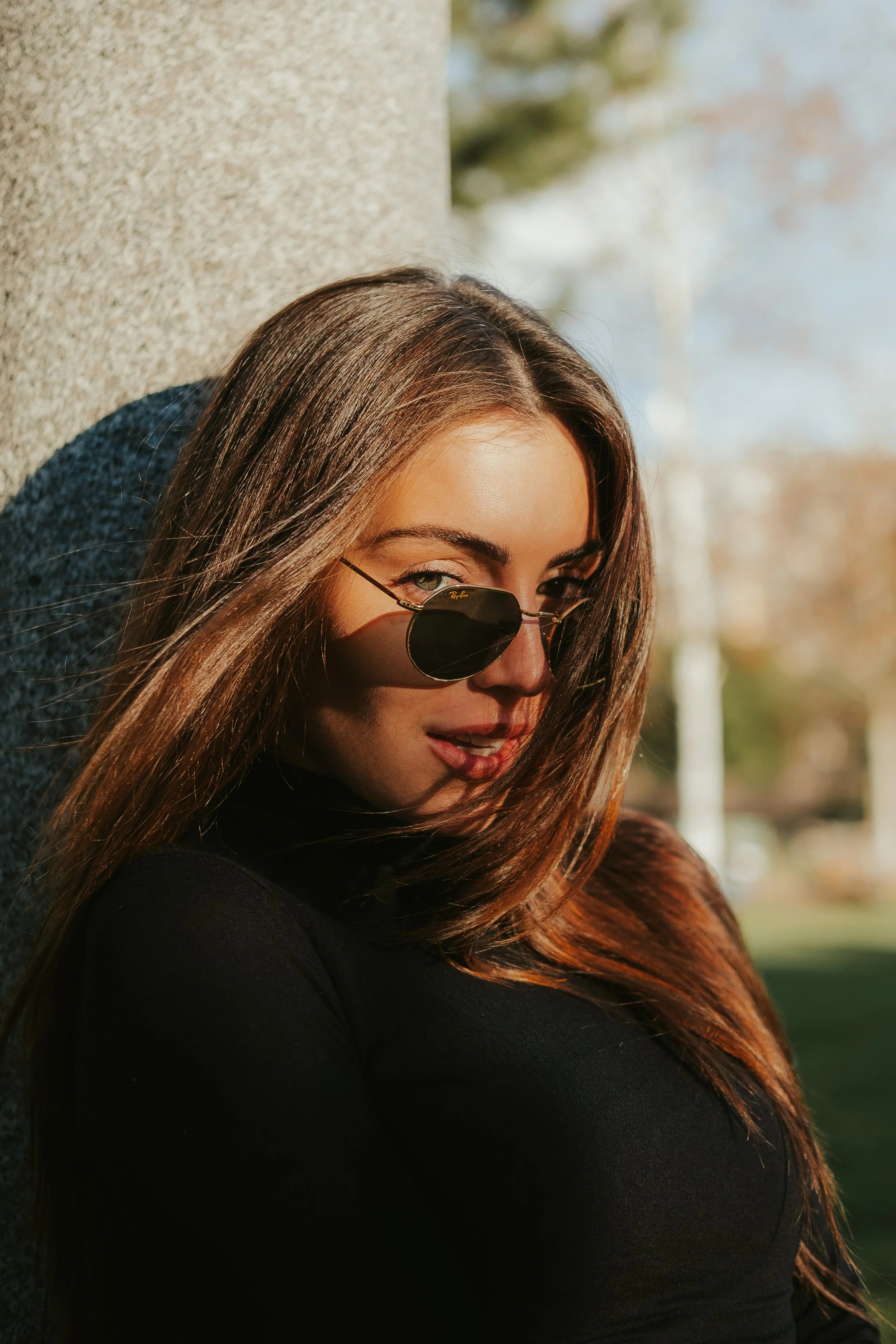 Mulher de cabelo castanho usando óculos escuros preto, vestindo roupa preta, apoiada em uma parede de concreto ao ar livre durante o dia.