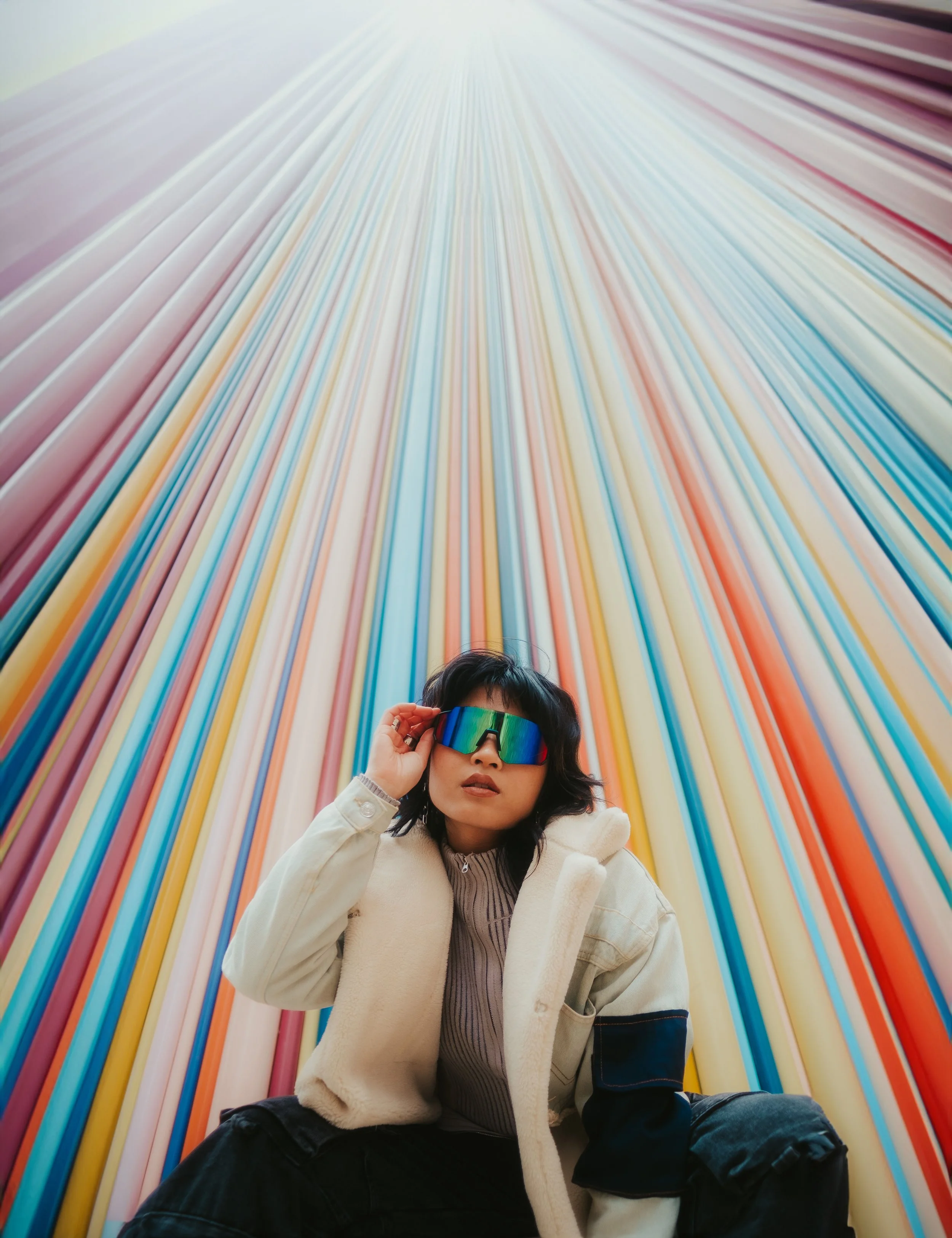 Pessoa com óculos de sol refletivos diante de parede com tubos de cores variadas, em um ambiente artístico e futurista.