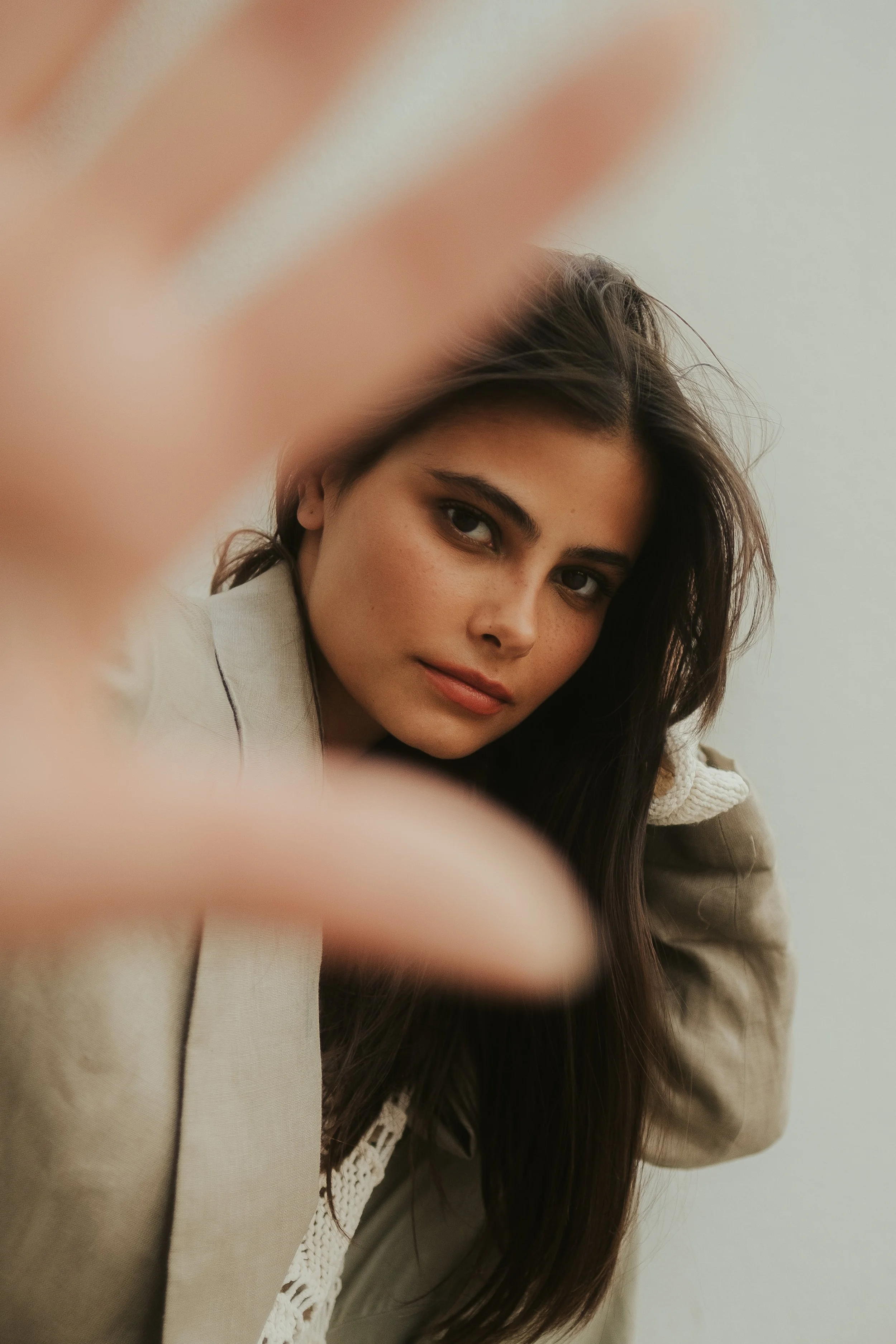 Jovem mulher com cabelo castanho escuro, usando jaqueta bege e camiseta branca, olhando para a câmera com expressão neutra, segurando a mão na frente parcialmente desfocada