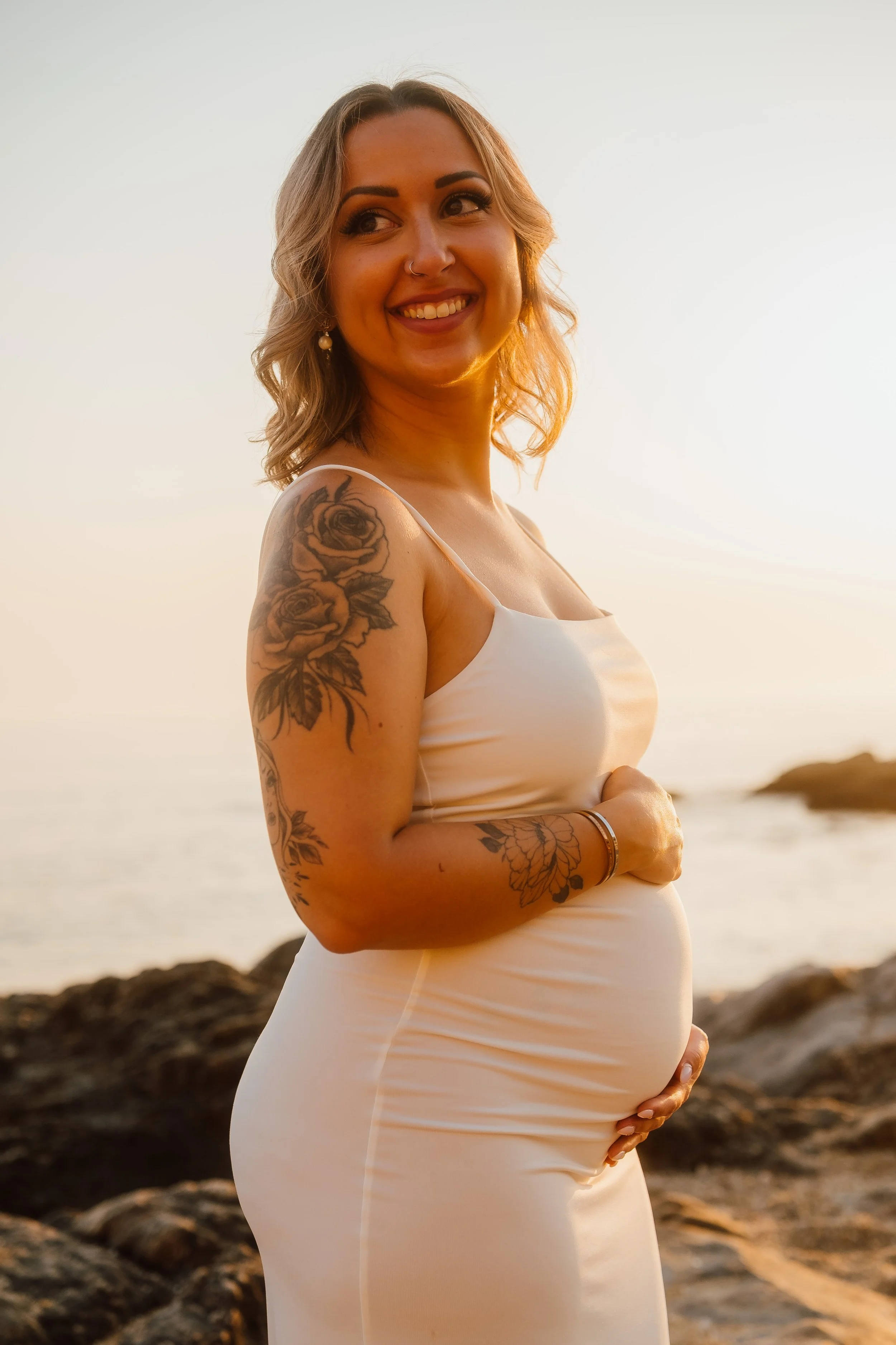 Mulher grávida com tatuagens, sorrindo na praia ao pôr do sol