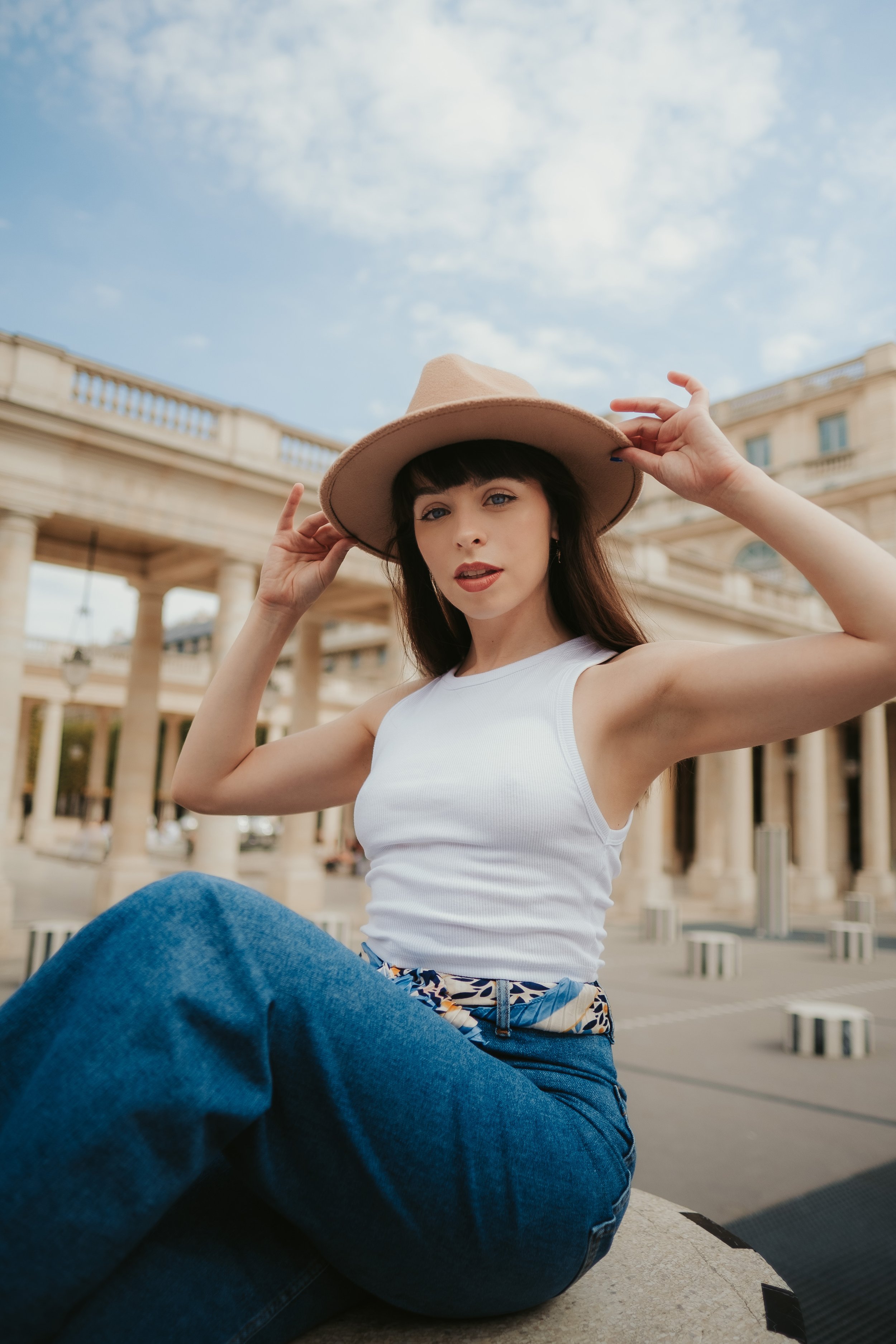 Jovem mulher com chapéu, camiseta branca, brincos, jeans azul e lenço na cintura, sentada ao ar livre com edifícios clássicos ao fundo.