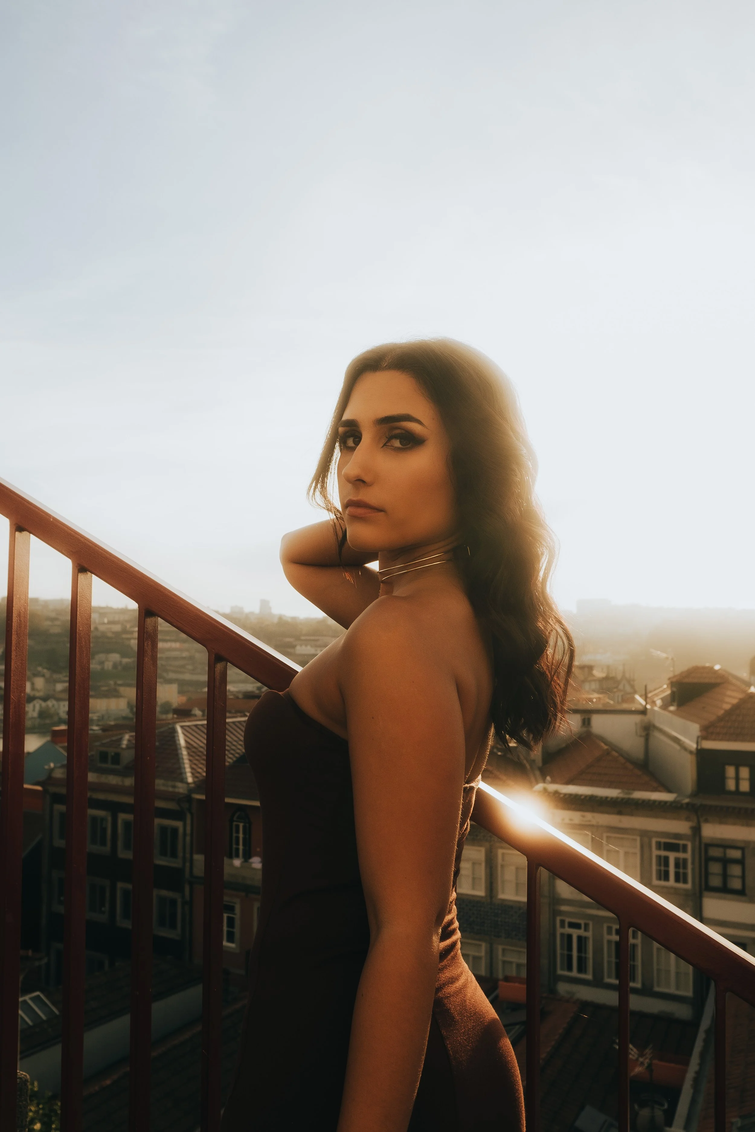 Mulher de cabelos cacheados e olhos marcantes posando na varanda ao pôr do sol, com vista da cidade ao fundo.