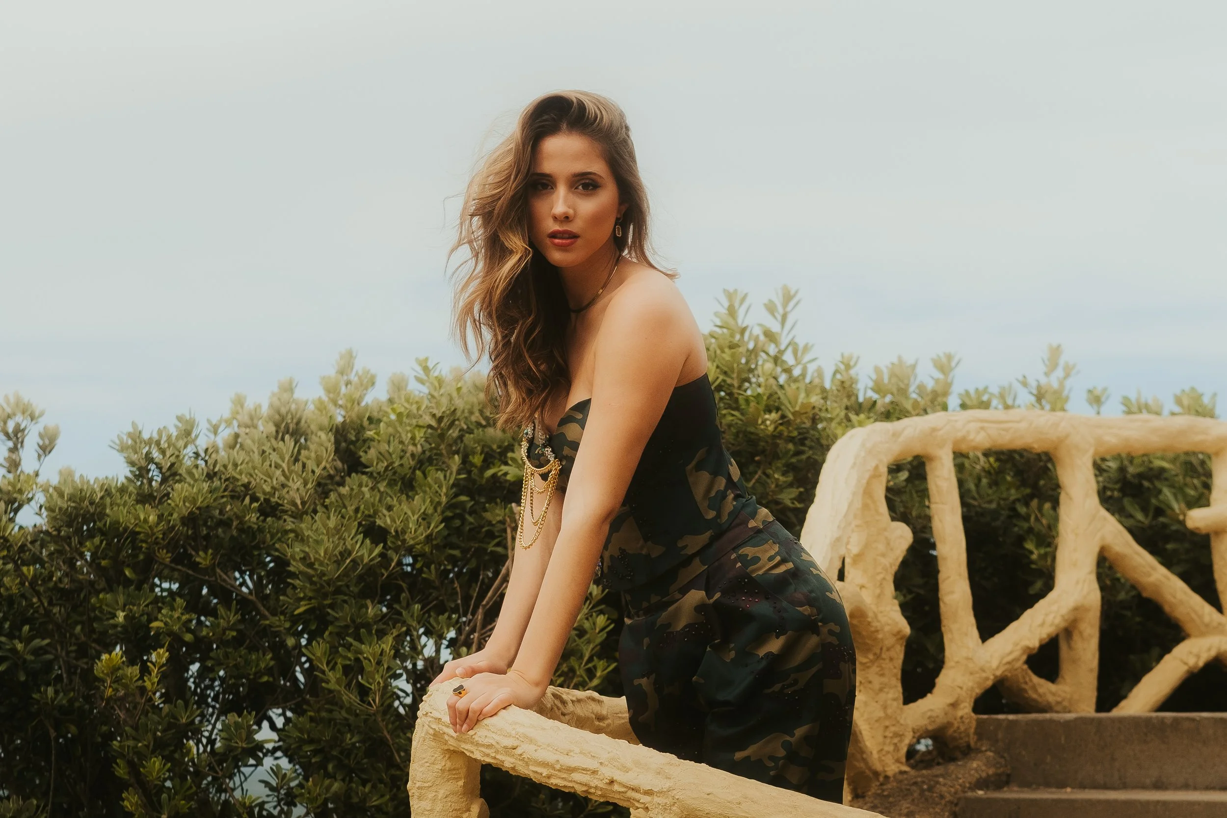 Mulher com cabelo castanho longo posando ao ar livre, usando roupa de camouflage, apoiada em uma grade de pedra, com vegetação ao fundo.
