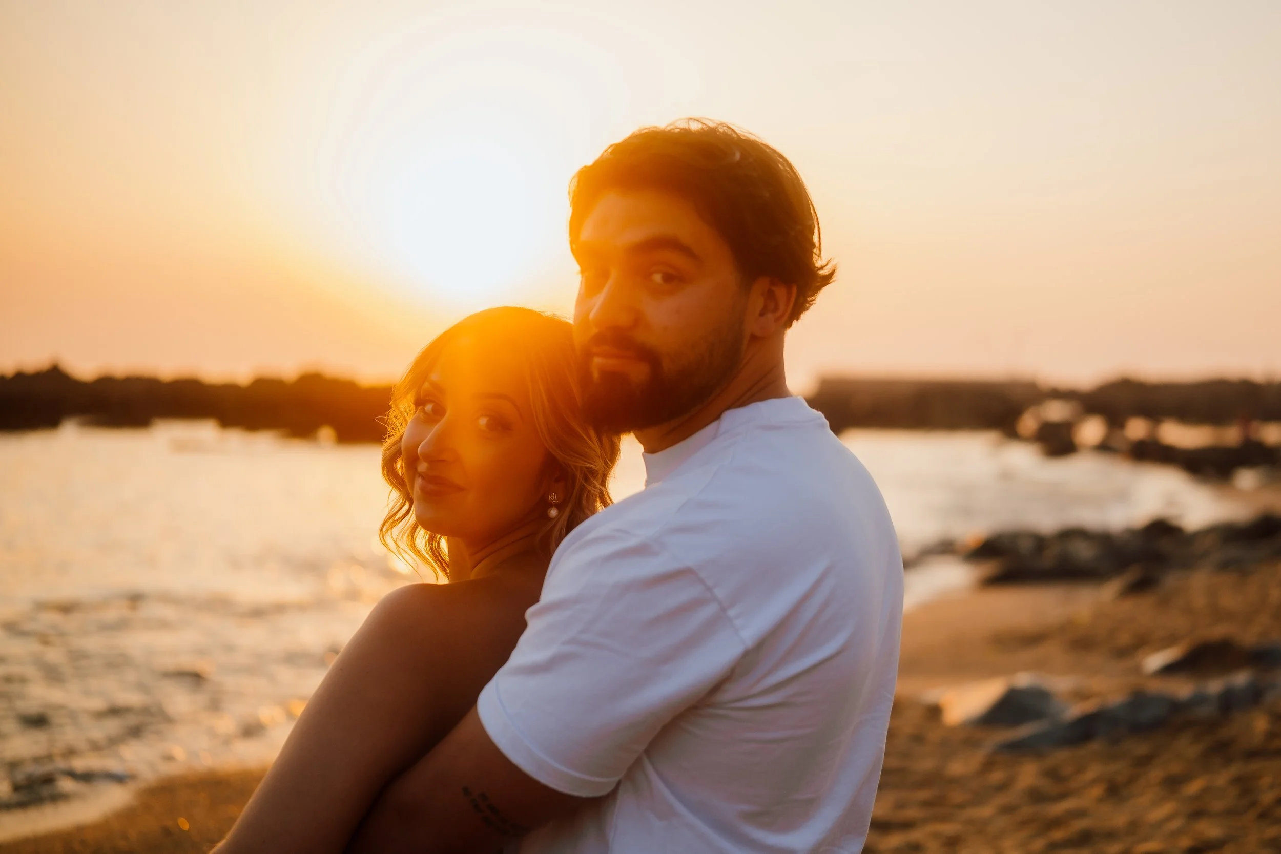 Um casal abraçado na praia ao pôr do sol, com o sol avermelhado no horizonte ao fundo.
