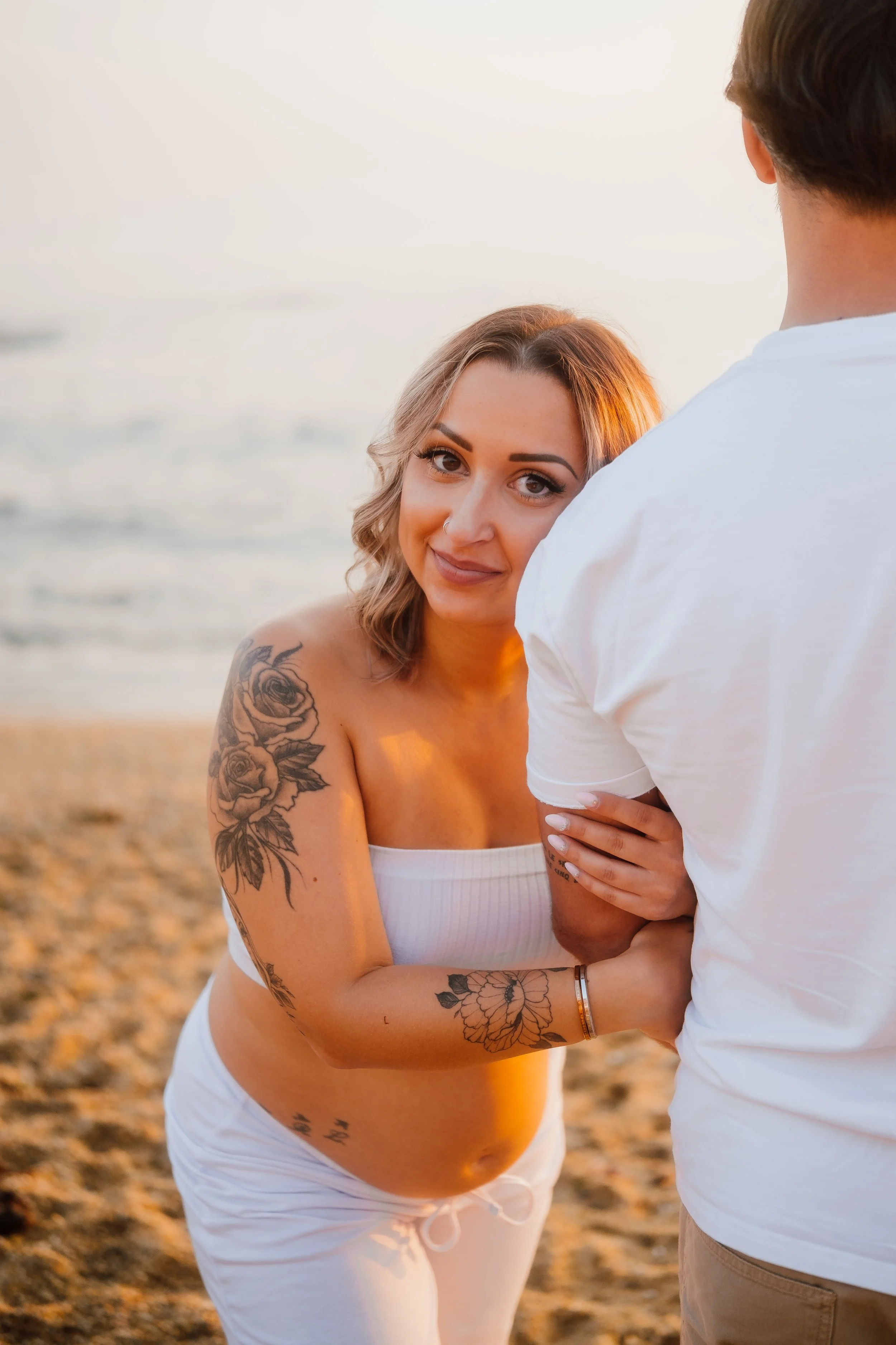 Casal na praia ao pôr do sol, mulher grávida com tatuagens no braço, usando roupa branca, segurando a mão de um homem com camiseta branca.