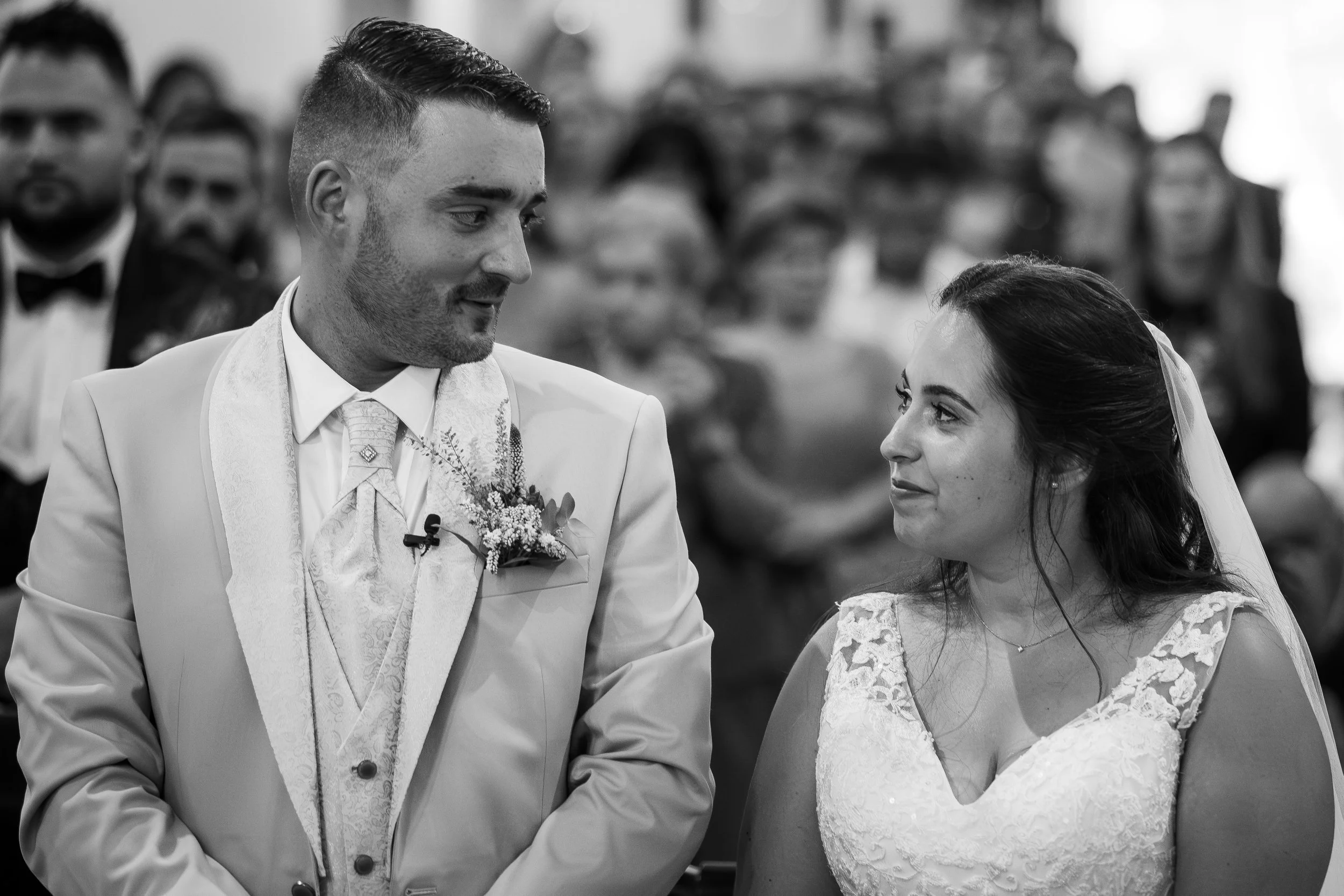 Casal de noivos em cerimônia de casamento, com convidados ao fundo.