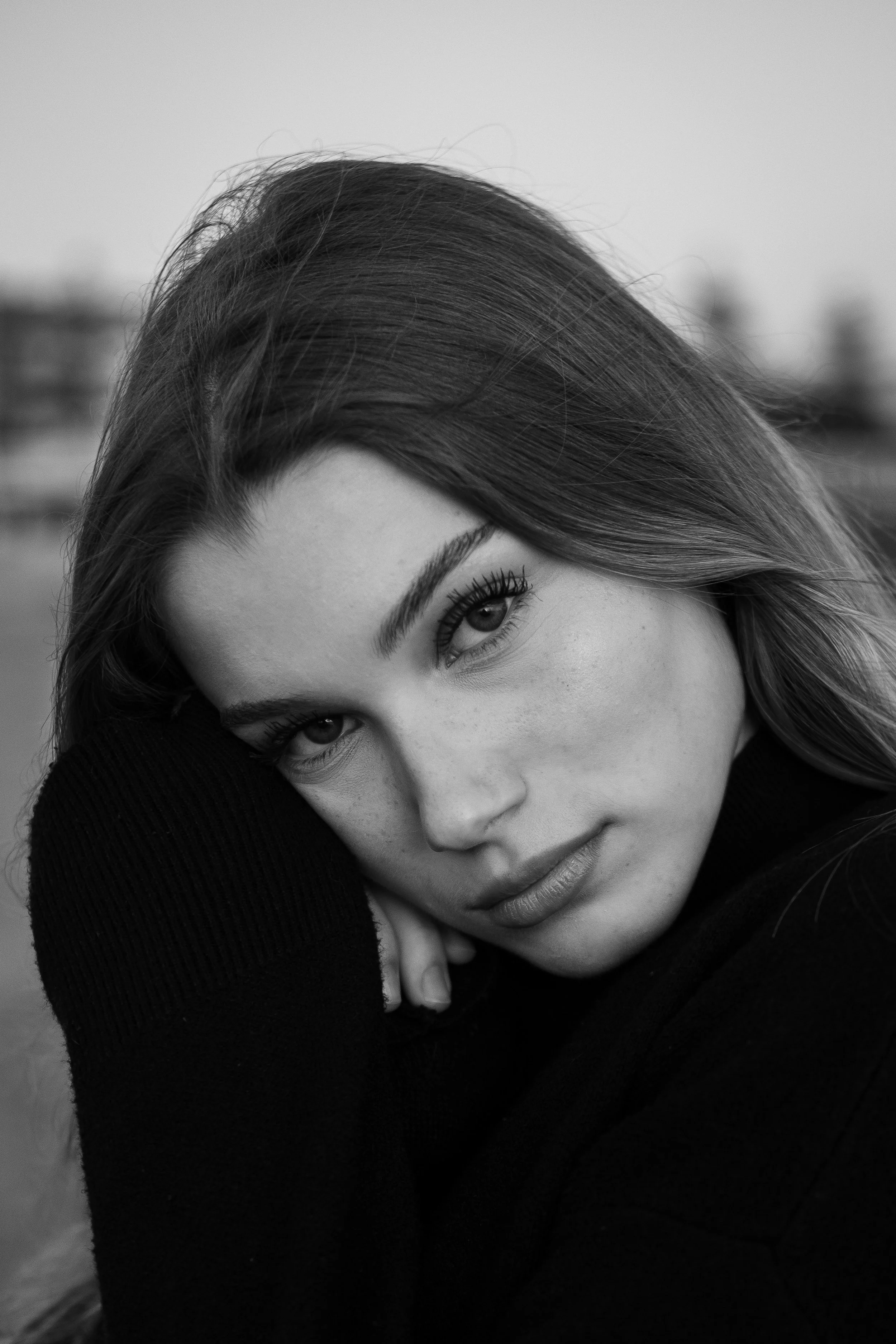 Retrato em preto e branco de uma mulher com cabelos longos, olhando para a câmera com expressão serena, apoiando a cabeça na mão, com fundo desfocado ao fundo.